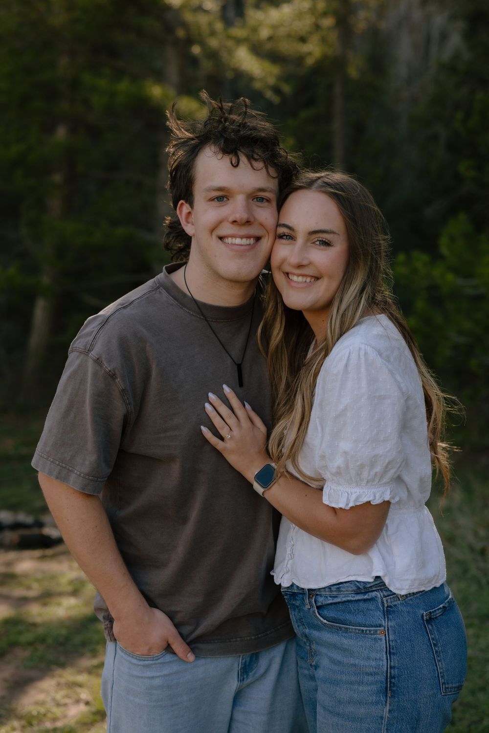 Happy engaged couple posing outdoors in forest, woman in white top and jeans hugging man in brown tee.