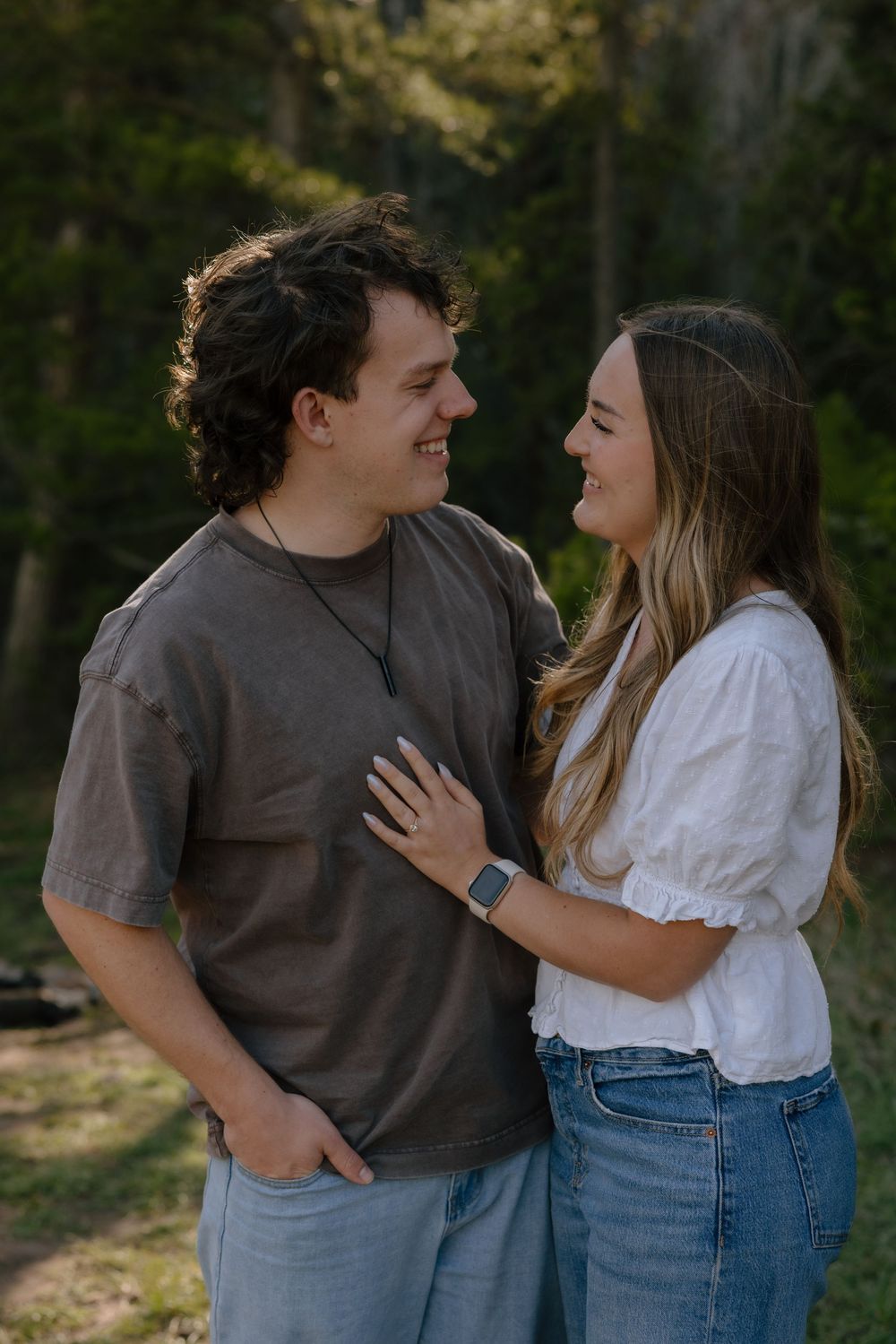 A smiling couple gazes at each other outdoors in a sunlit forest setting.