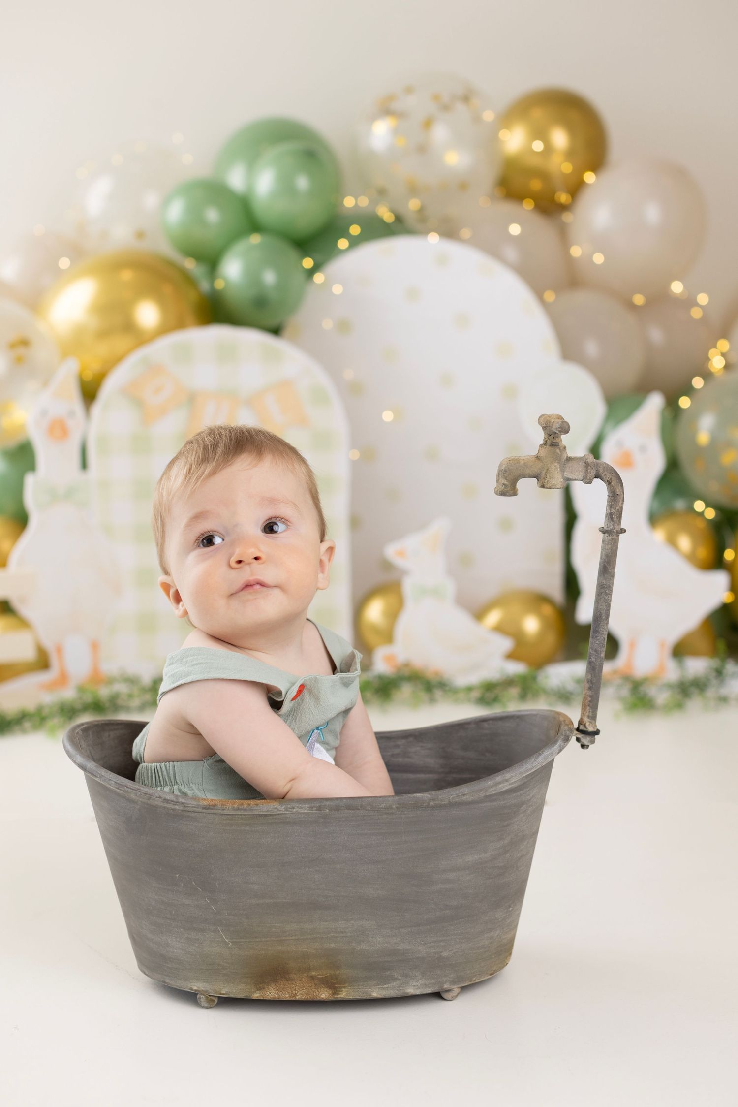 Baby in vintage tub with green and gold balloon backdrop for a first birthday photo session.