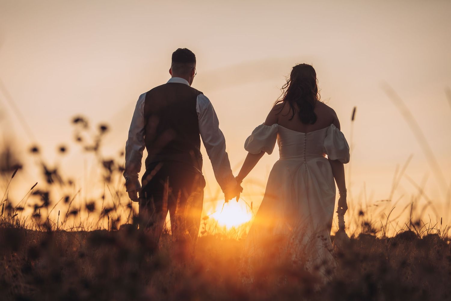 Silhouette of a couple holding hands in a golden sunset field, romantic wedding photo with warm backlit glow.
