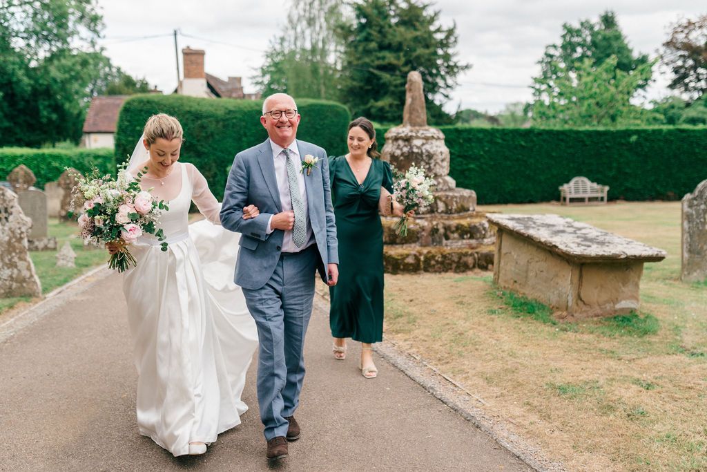 Bride in white gown walks with family through a churchyard on her wedding day, holding a bouquet.