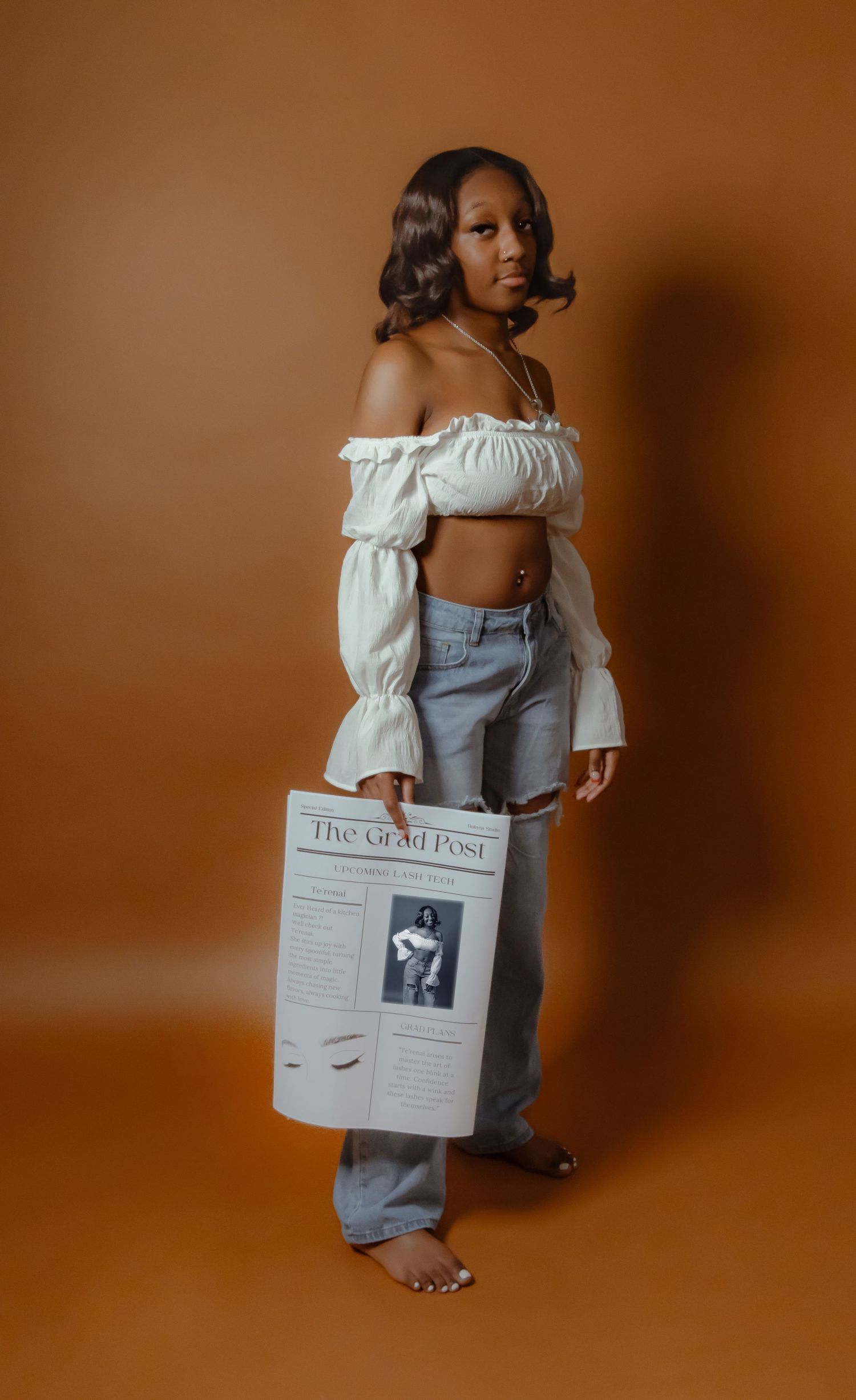 Young woman in white off-shoulder crop top and ripped jeans holding The Grad Post newspaper against brown backdrop.