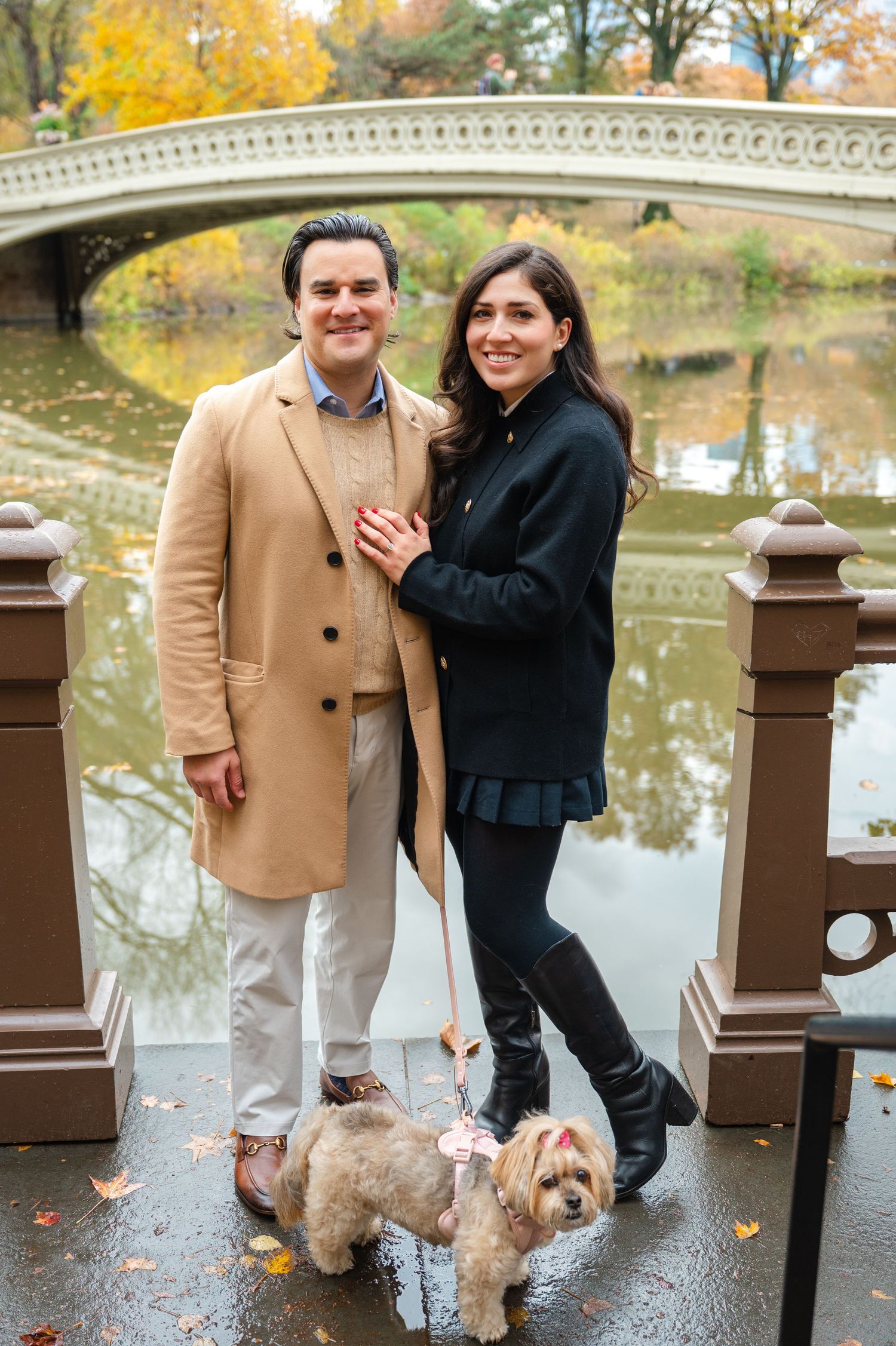 Couple posing with small dog on leash near Bow Bridge in Central Park during autumn, wearing elegant coats after a proposal session in NYC.