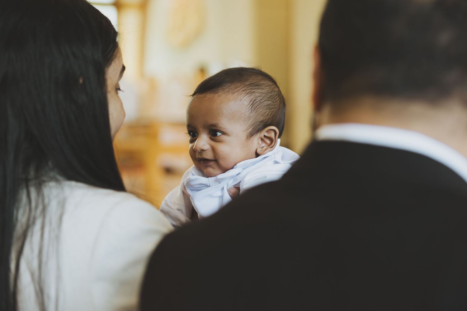 Smiling baby in white outfit being held by parents during a christening ceremony.