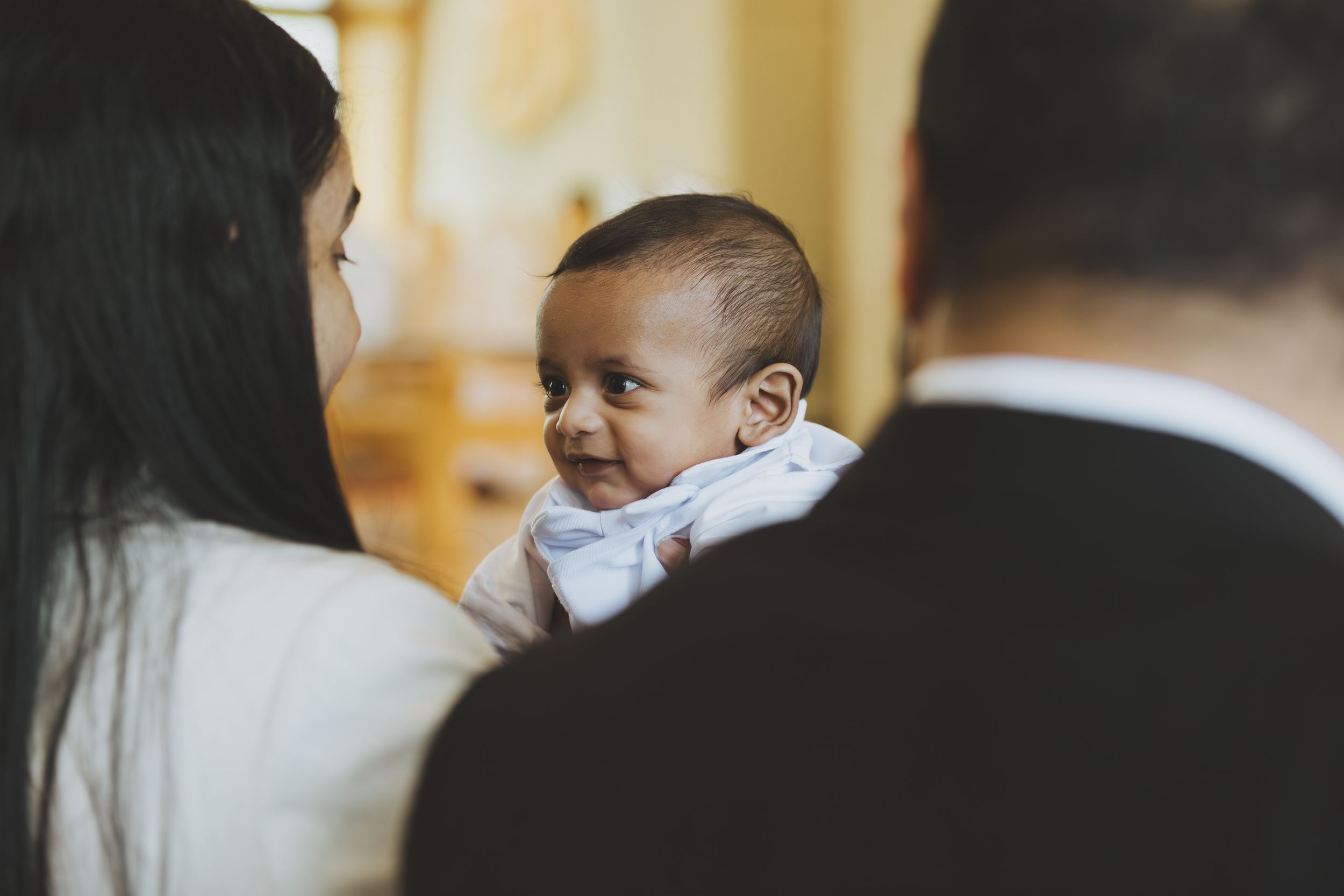 Smiling baby in white outfit being held by parents during a christening ceremony.