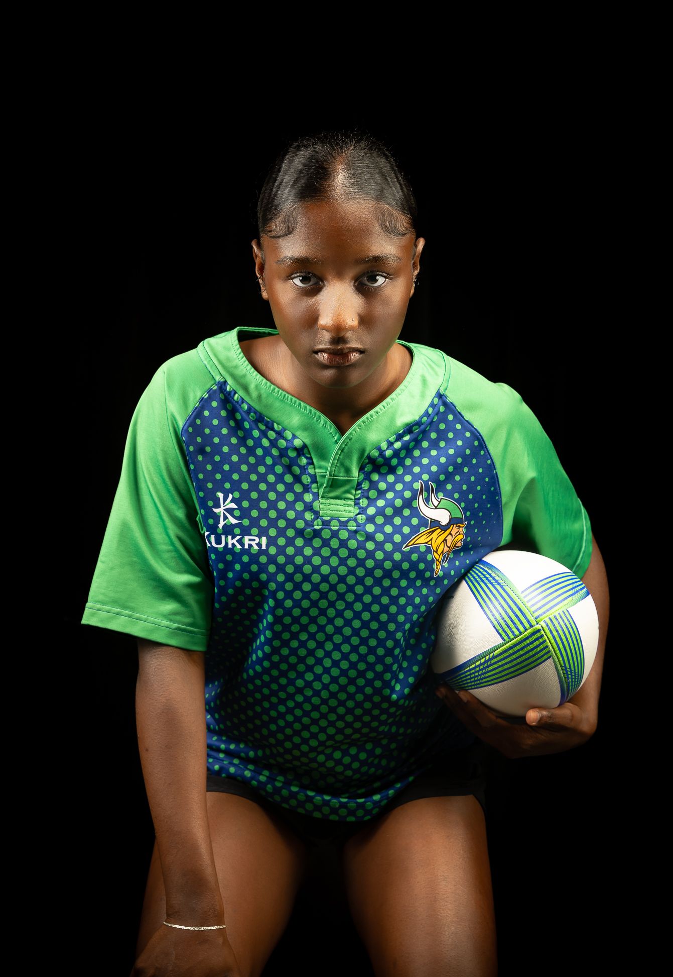 A girl in a green and blue rugby jersey, bending down and looking at the camera with a rugby ball under her left arm