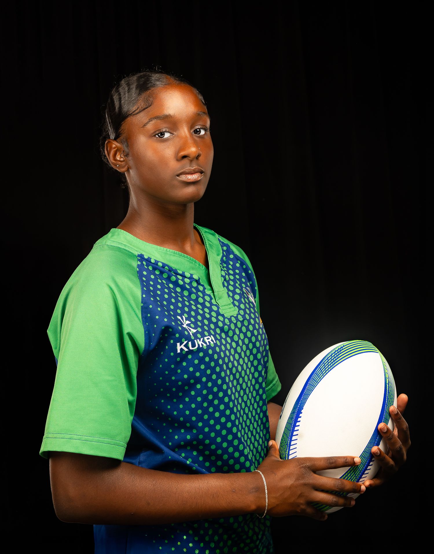 A girl in a green and blue rugby jersey, holding a rugby ball with body angled to the side
