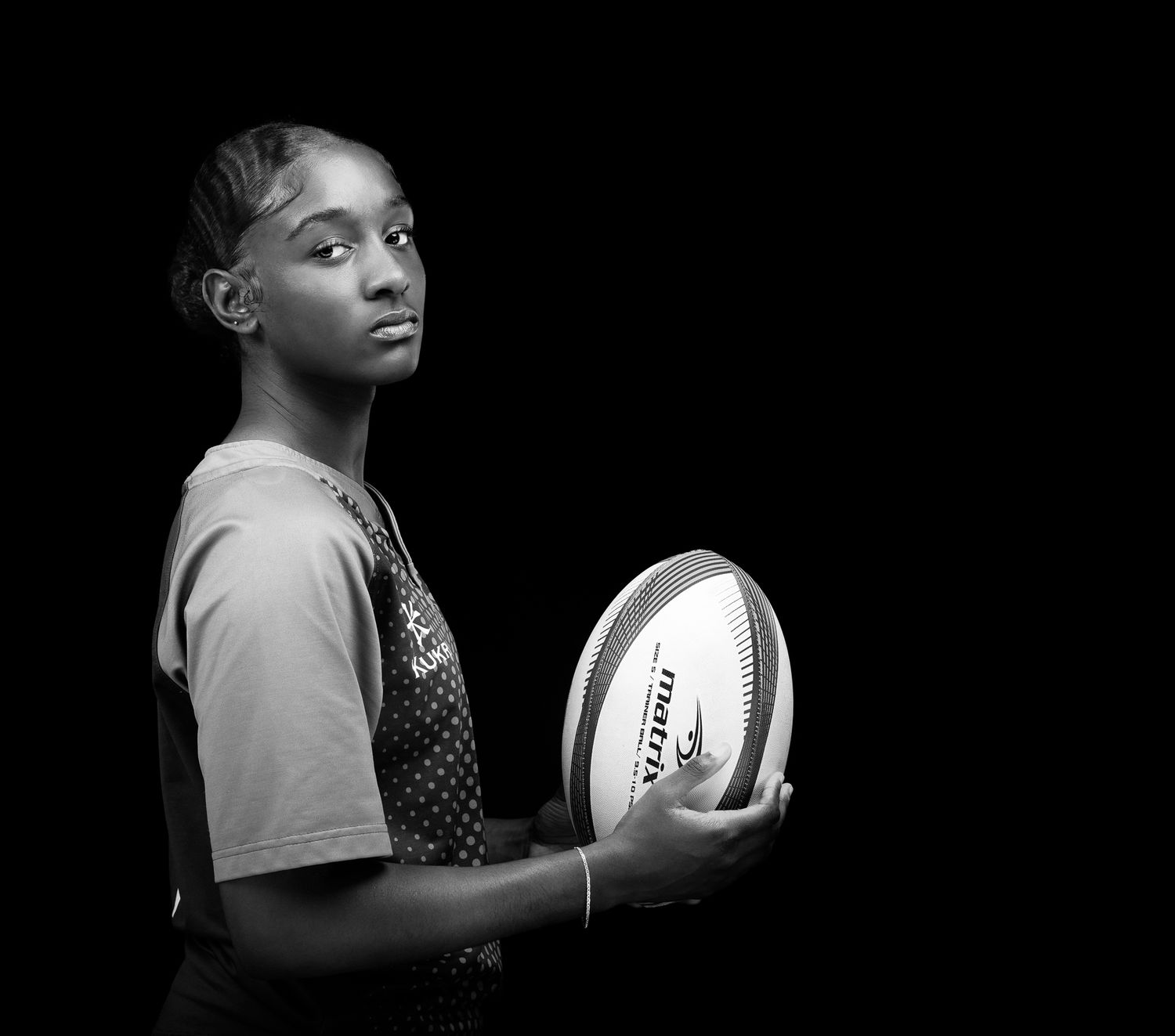 A black and white image of a girl holding a rugby ball with her body facing the right & head facing straight to the camera
