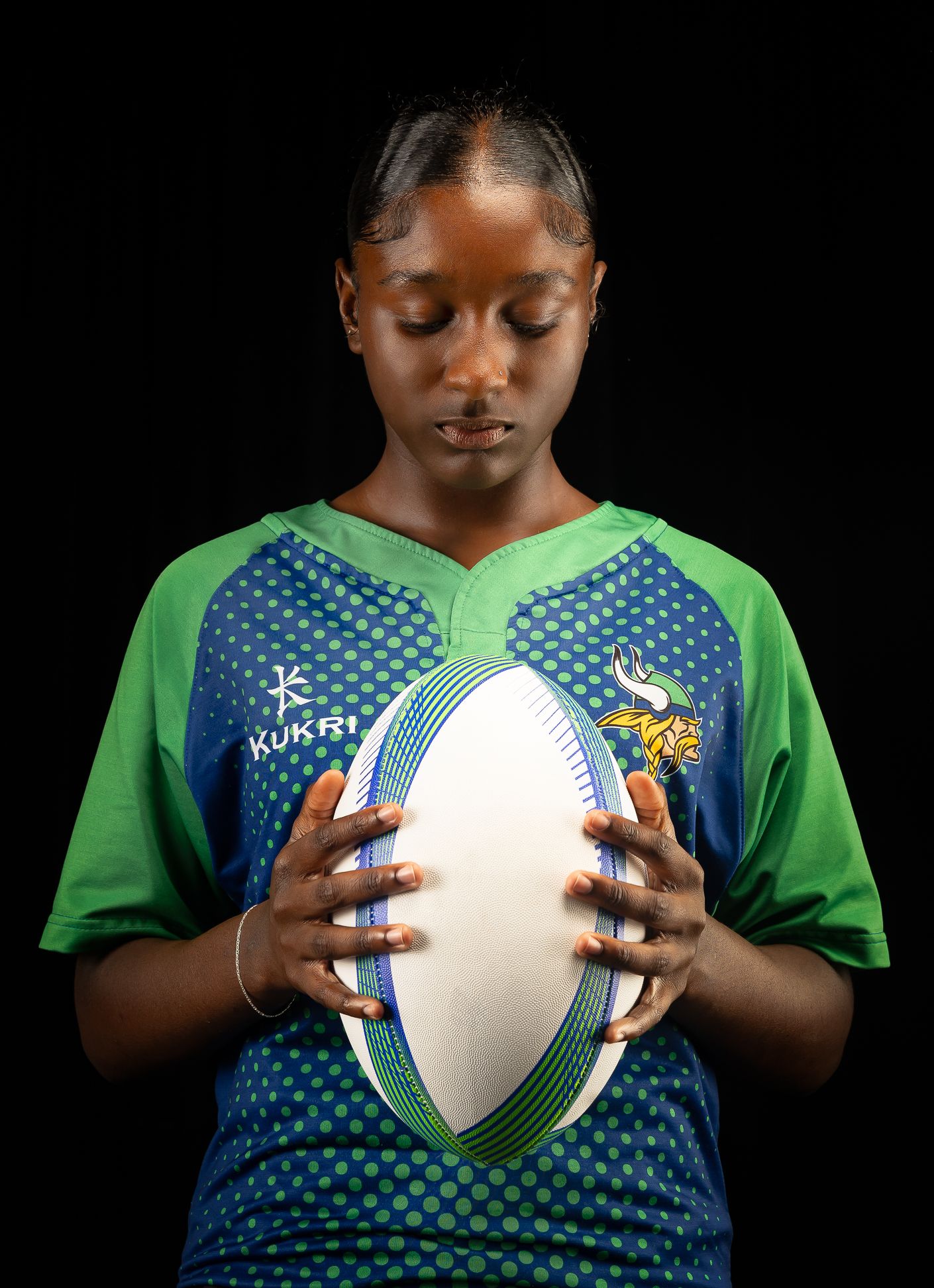 An image of a girl in a green and blue rugby jersey, holding a white rugby ball looking down at it
