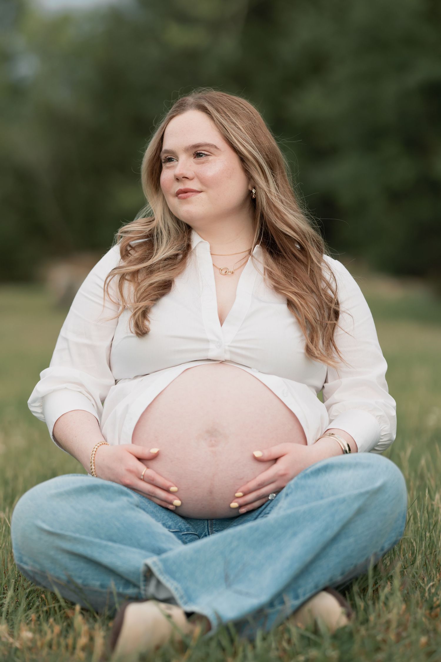 Pregnant woman sitting cross-legged on grass, cradling her baby bump, wearing white shirt and jeans outdoors.