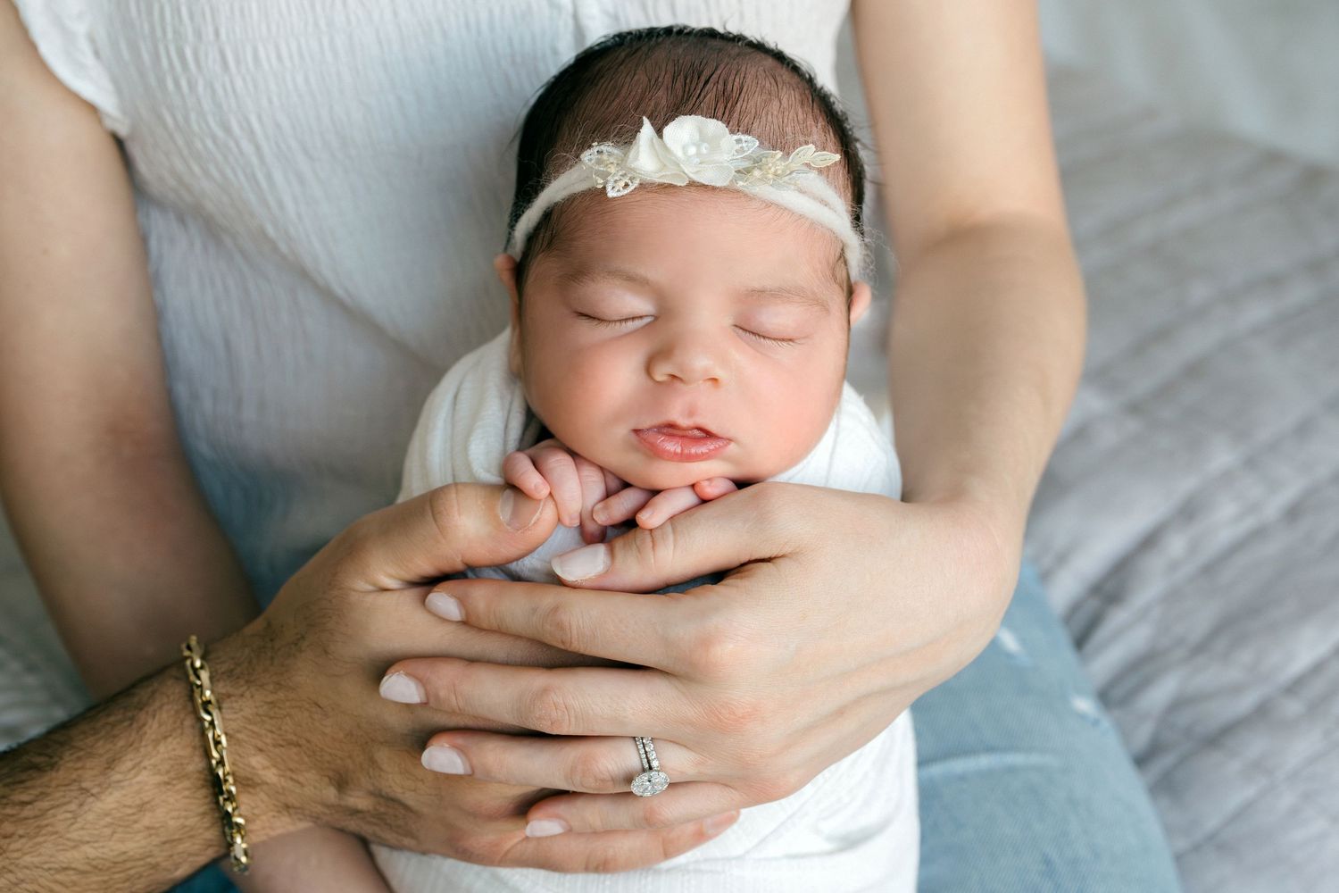 Sleeping newborn baby in white outfit and floral headband cradled in parents hands.