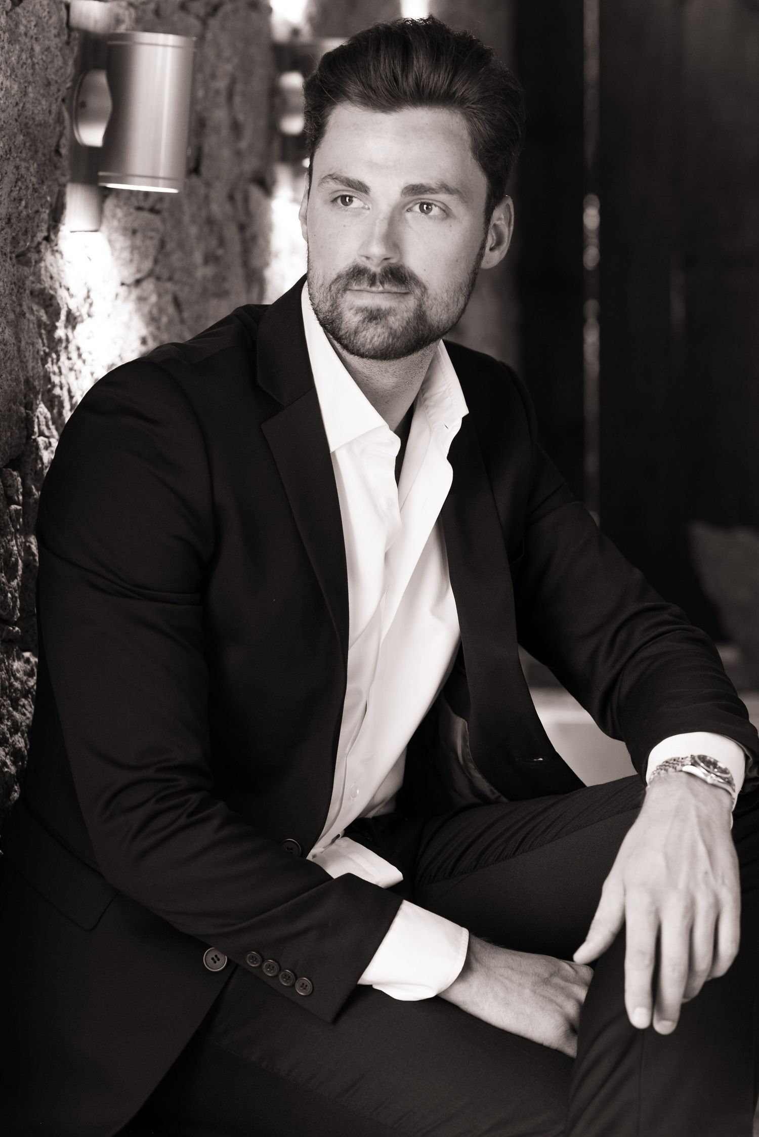 Black and white portrait of a bearded man in a black suit and white shirt sitting against a stone wall.