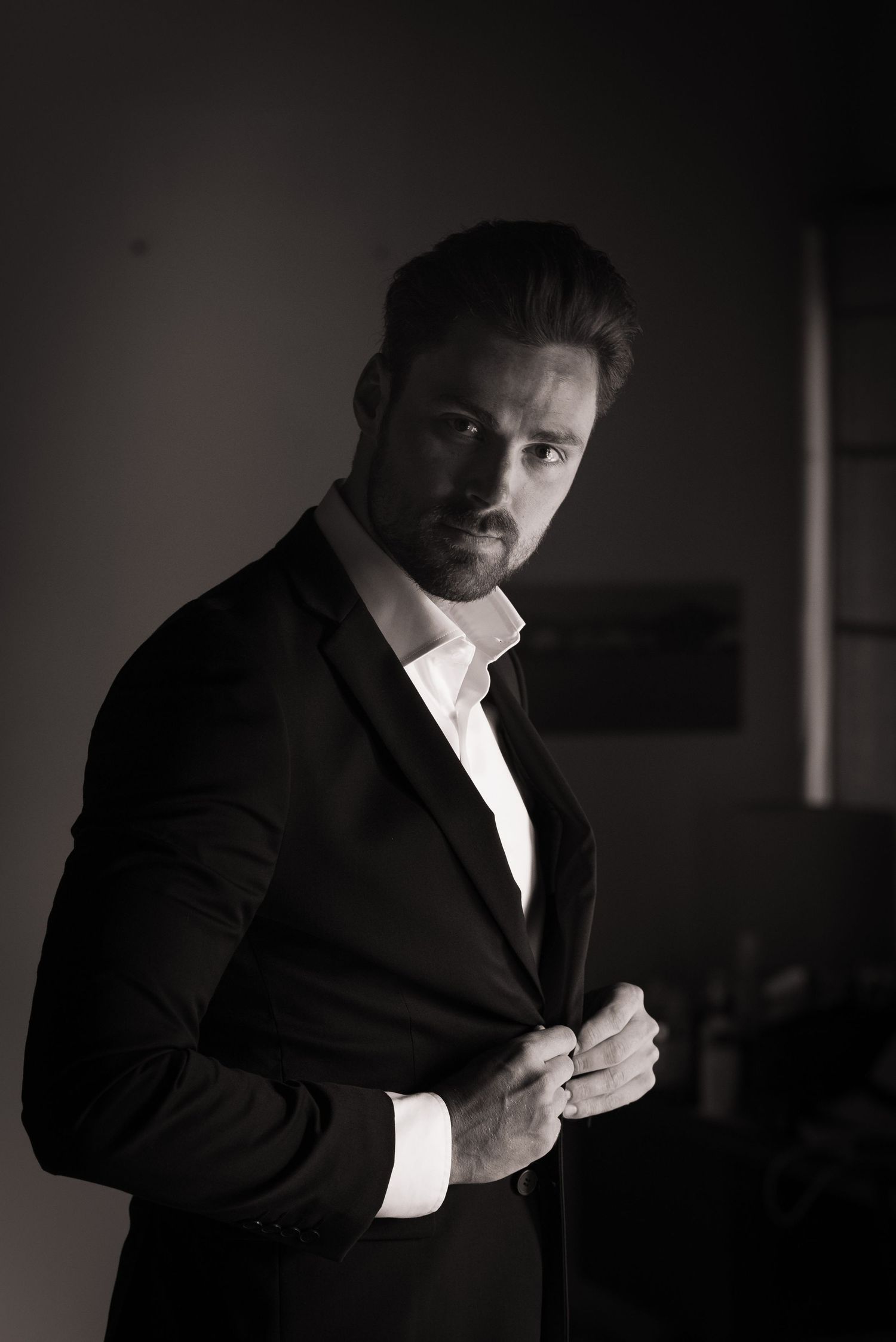 Black and white portrait of a well-dressed man in a dark suit buttoning his jacket indoors.