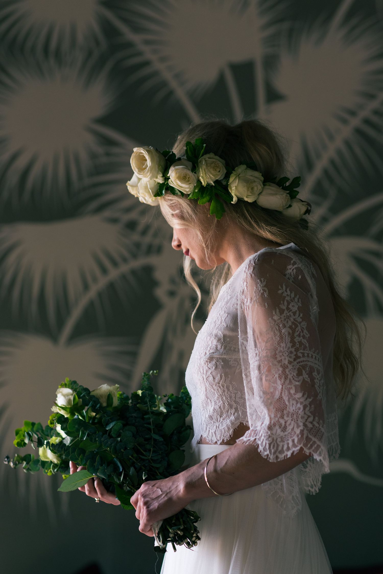 Bride in lace dress holding white rose bouquet, wearing floral crown, standing against tropical wallpaper.