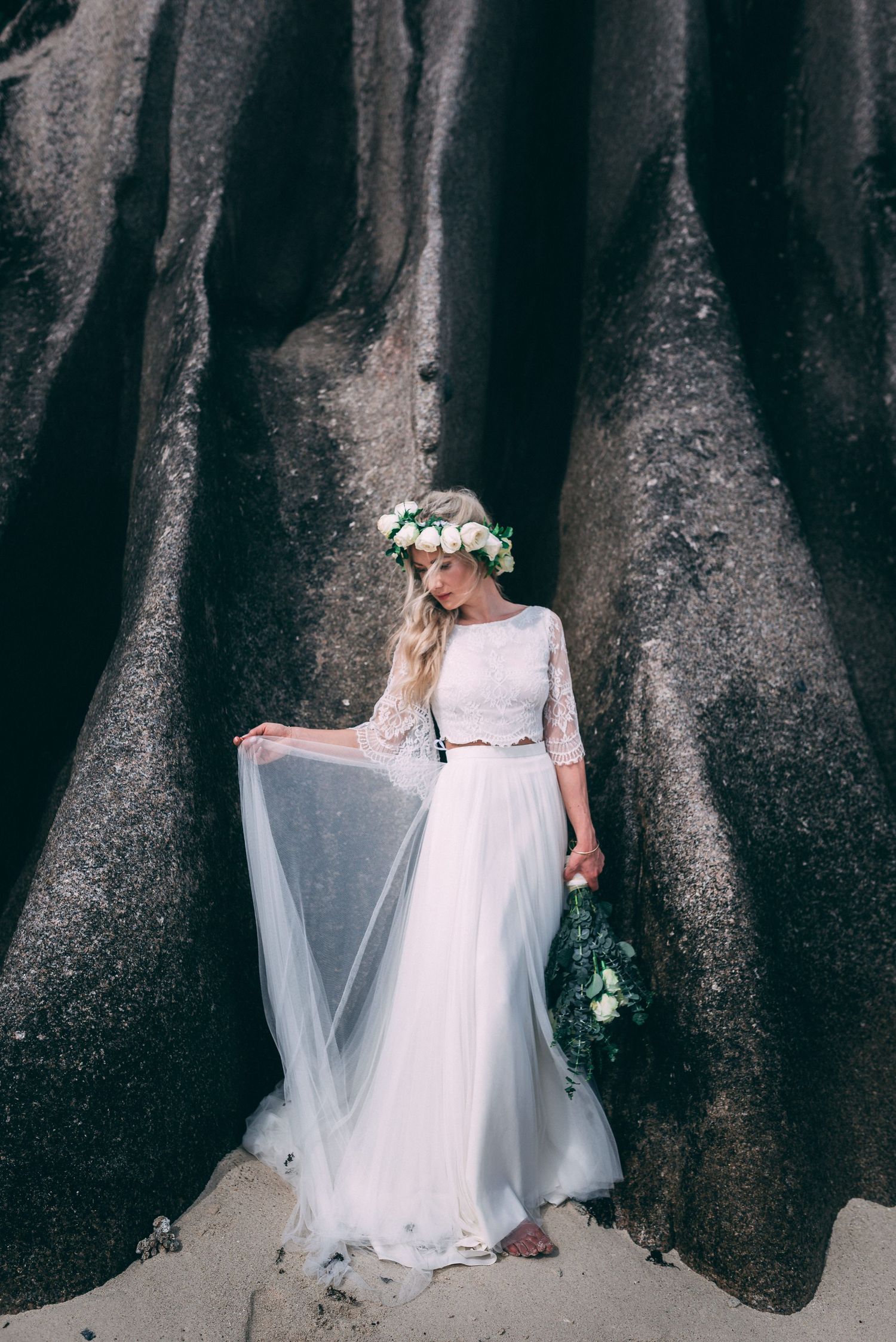 Bride in white lace gown and floral crown holds bouquet beside large tree roots on sandy ground.