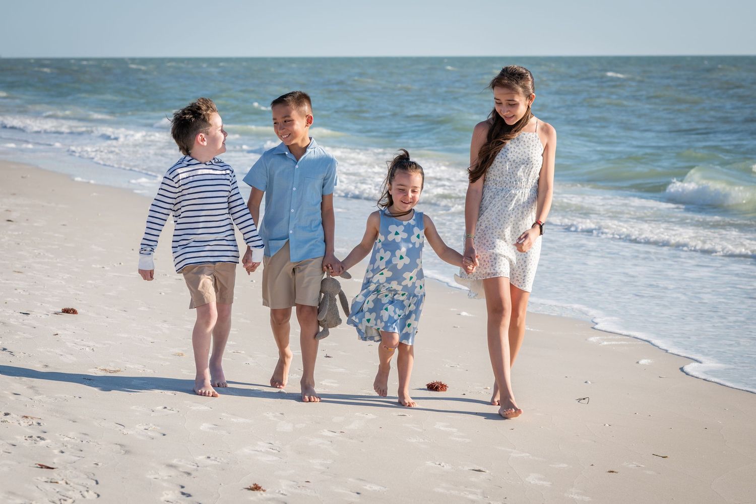 Four children walking hand-in-hand on a sunny beach with ocean waves in the background.