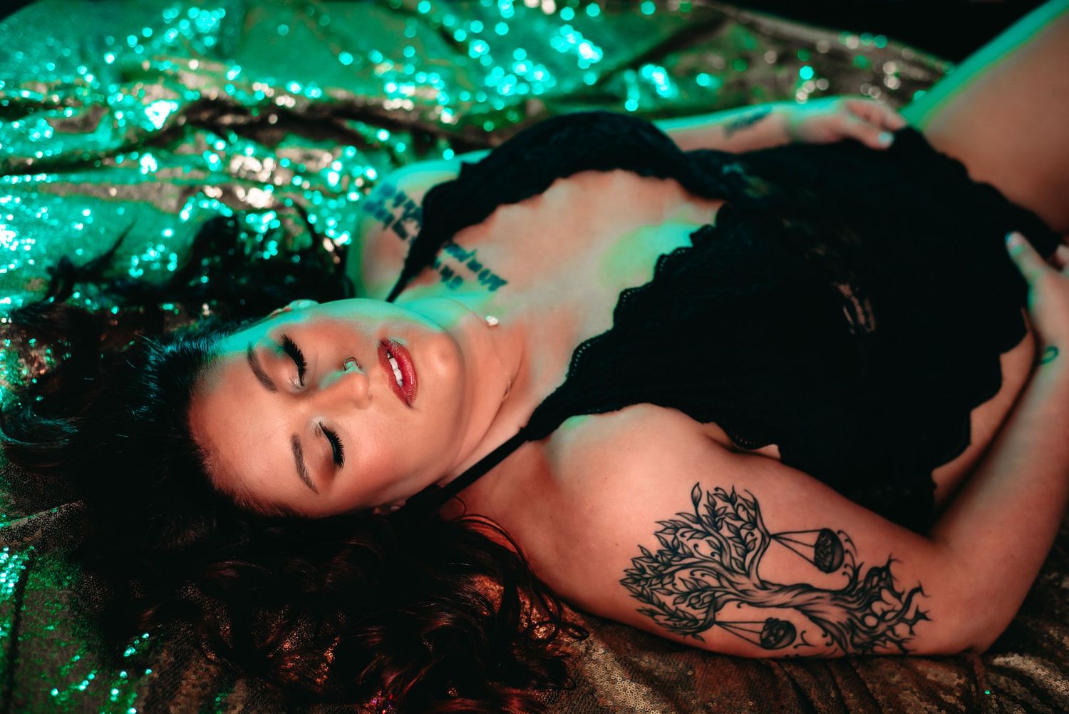 Smiling woman with curly hair lying on leopard print fabric, wearing red polka dot top with tattoos visible.