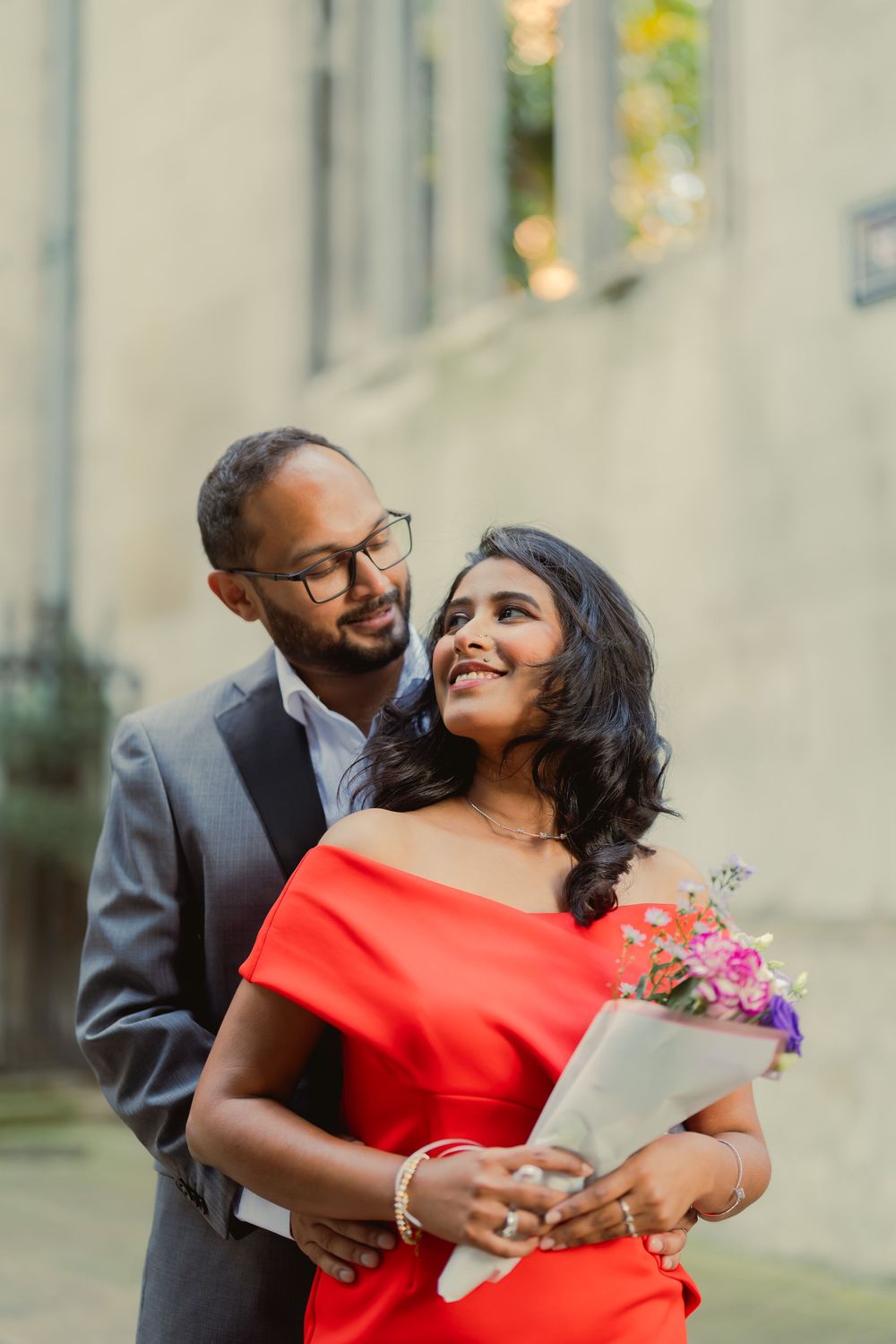 Couple in elegant attire embracing outdoors, woman in red off-shoulder dress holding flowers, man in gray suit with glasses.