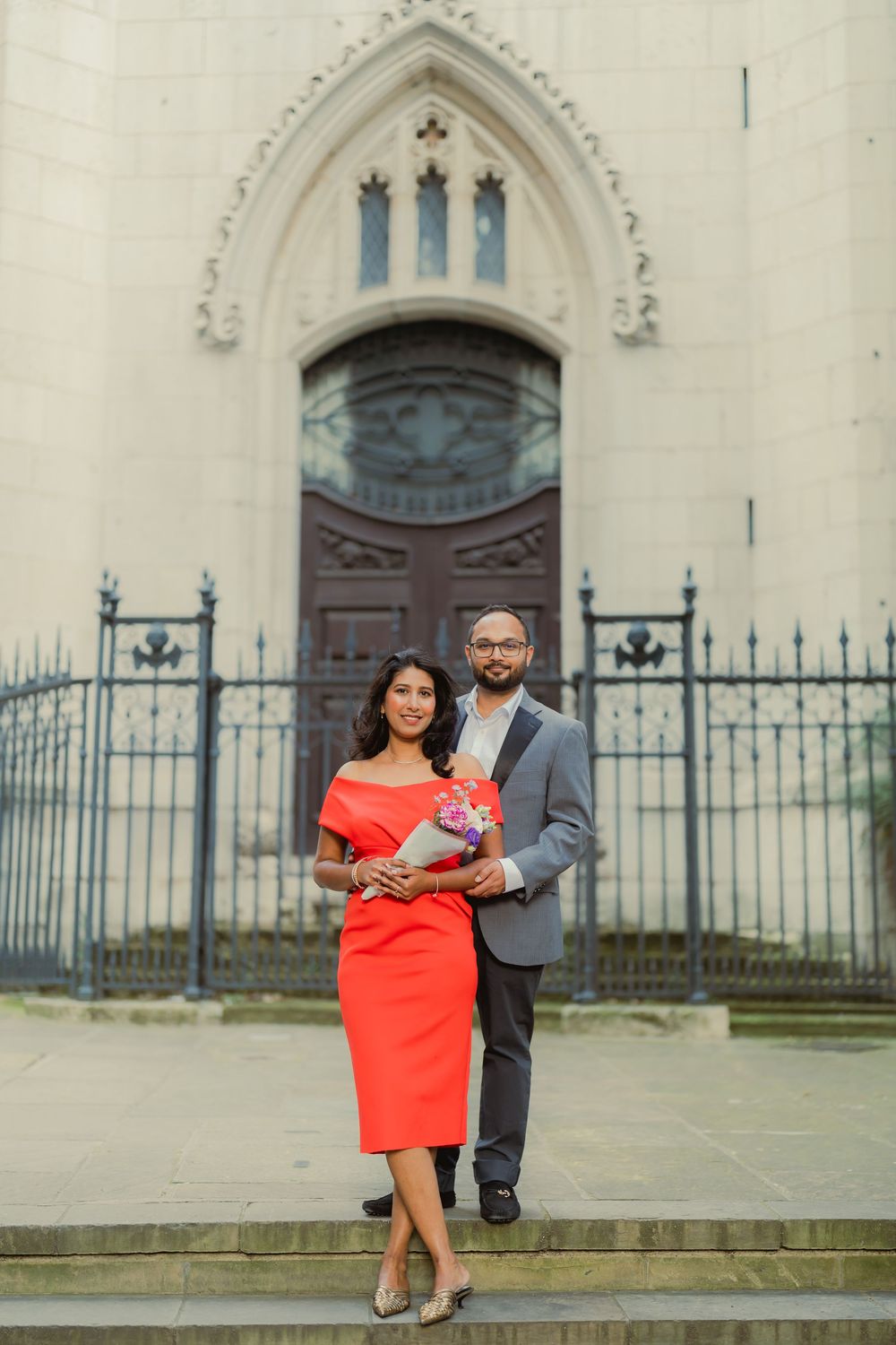 Couple posing in front of a Gothic church, woman in red dress holding bouquet, man in gray suit.