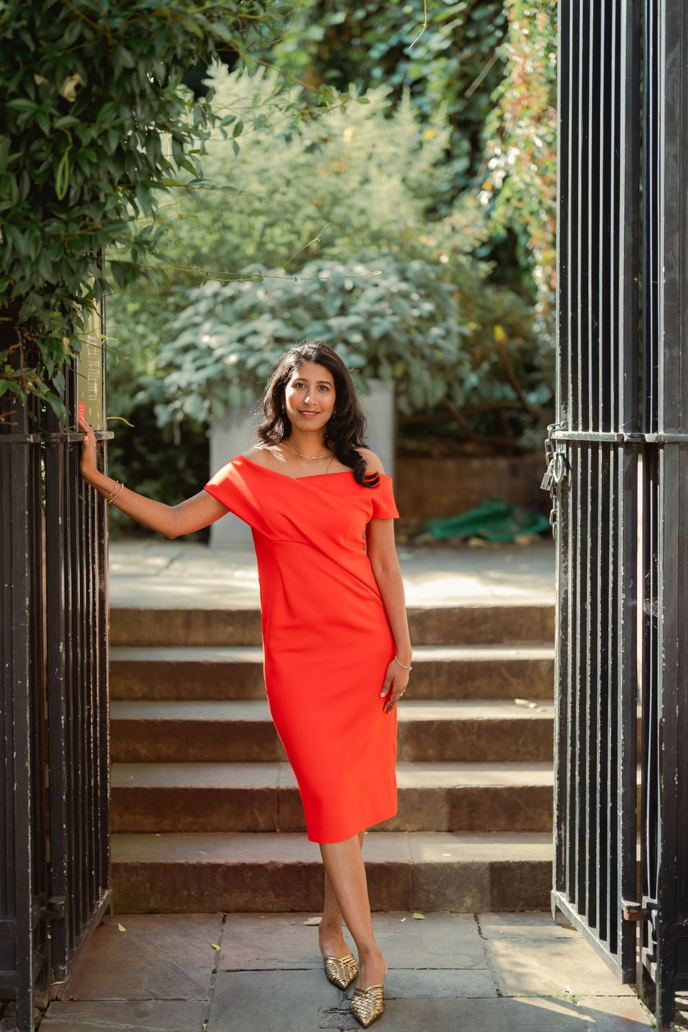 Woman in red off-shoulder midi dress posing at iron gate with lush greenery and stone steps background.