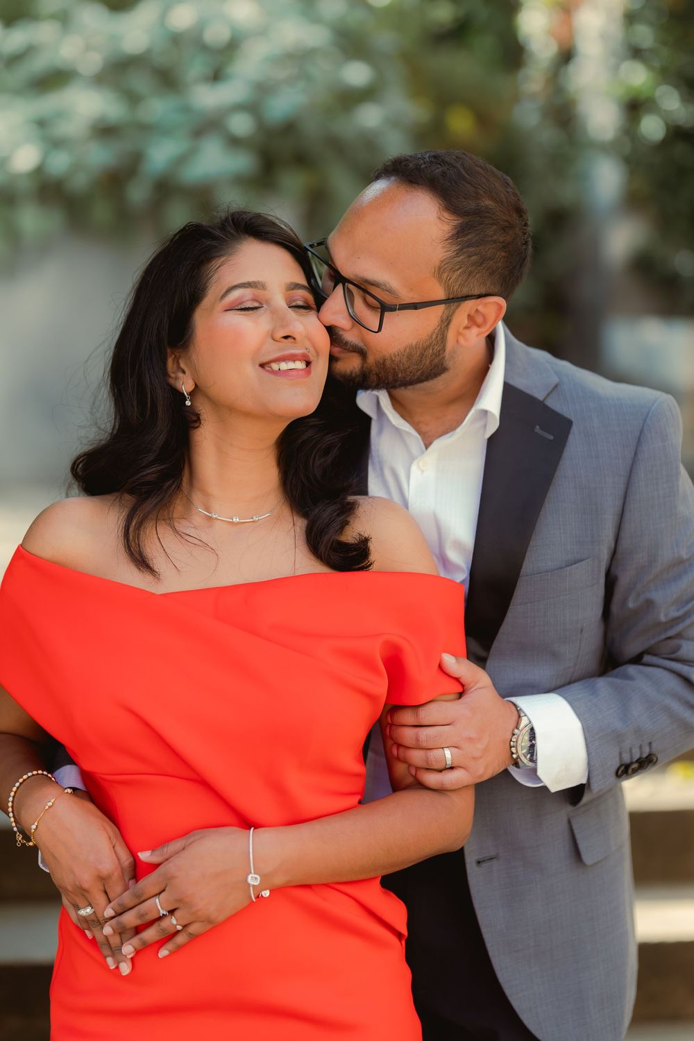 A couple shares a romantic moment, man in gray suit kisses woman in red off-shoulder dress outdoors.