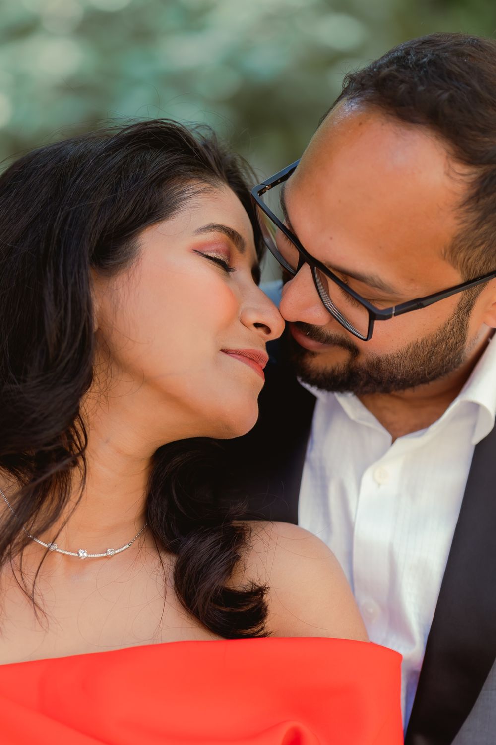 A romantic couple sharing an intimate moment, woman in red dress with silver necklace, man in glasses and white shirt.