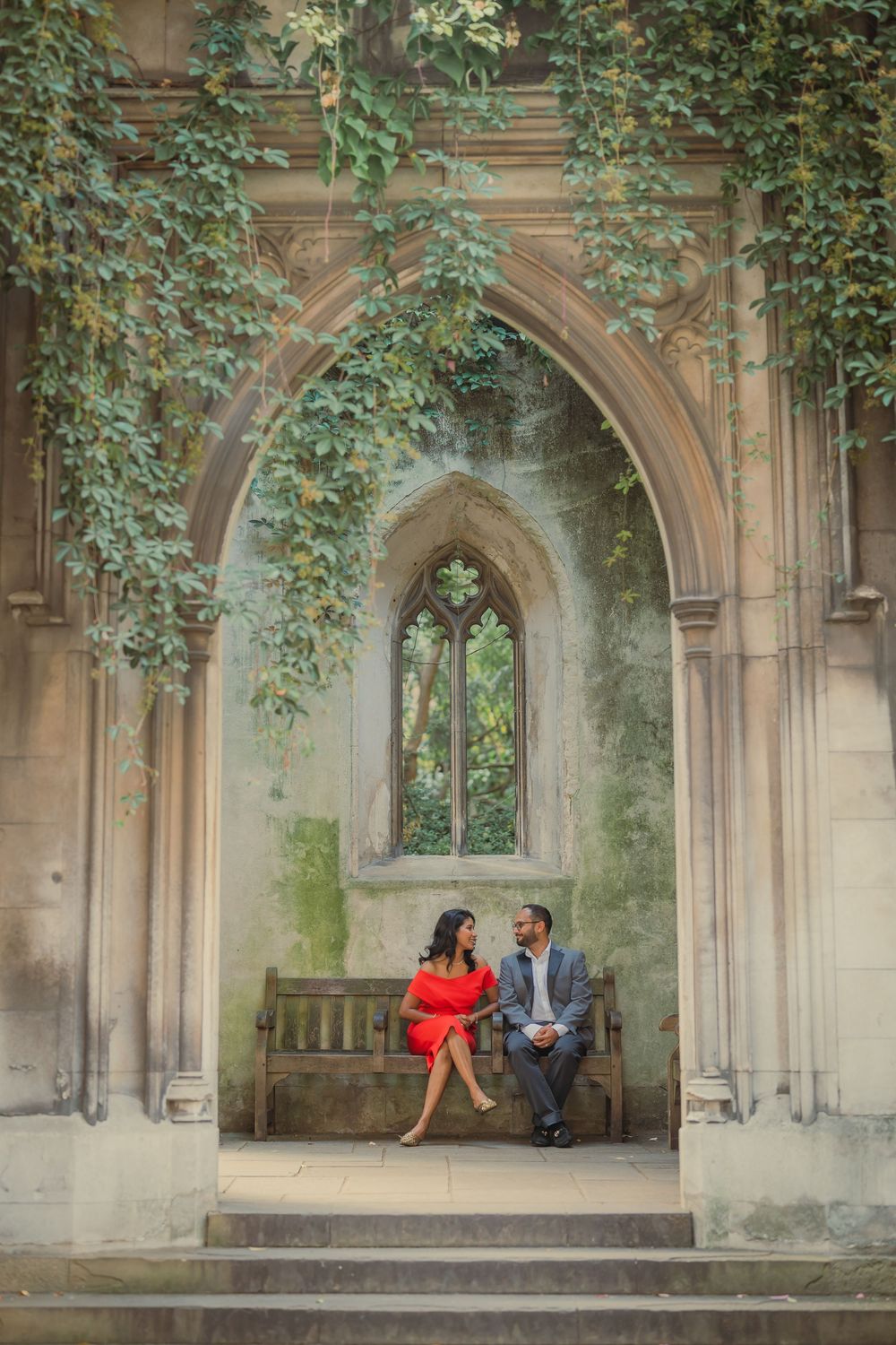 Couple sitting on a bench under a Gothic stone arch covered in ivy, woman in red dress, man in suit.