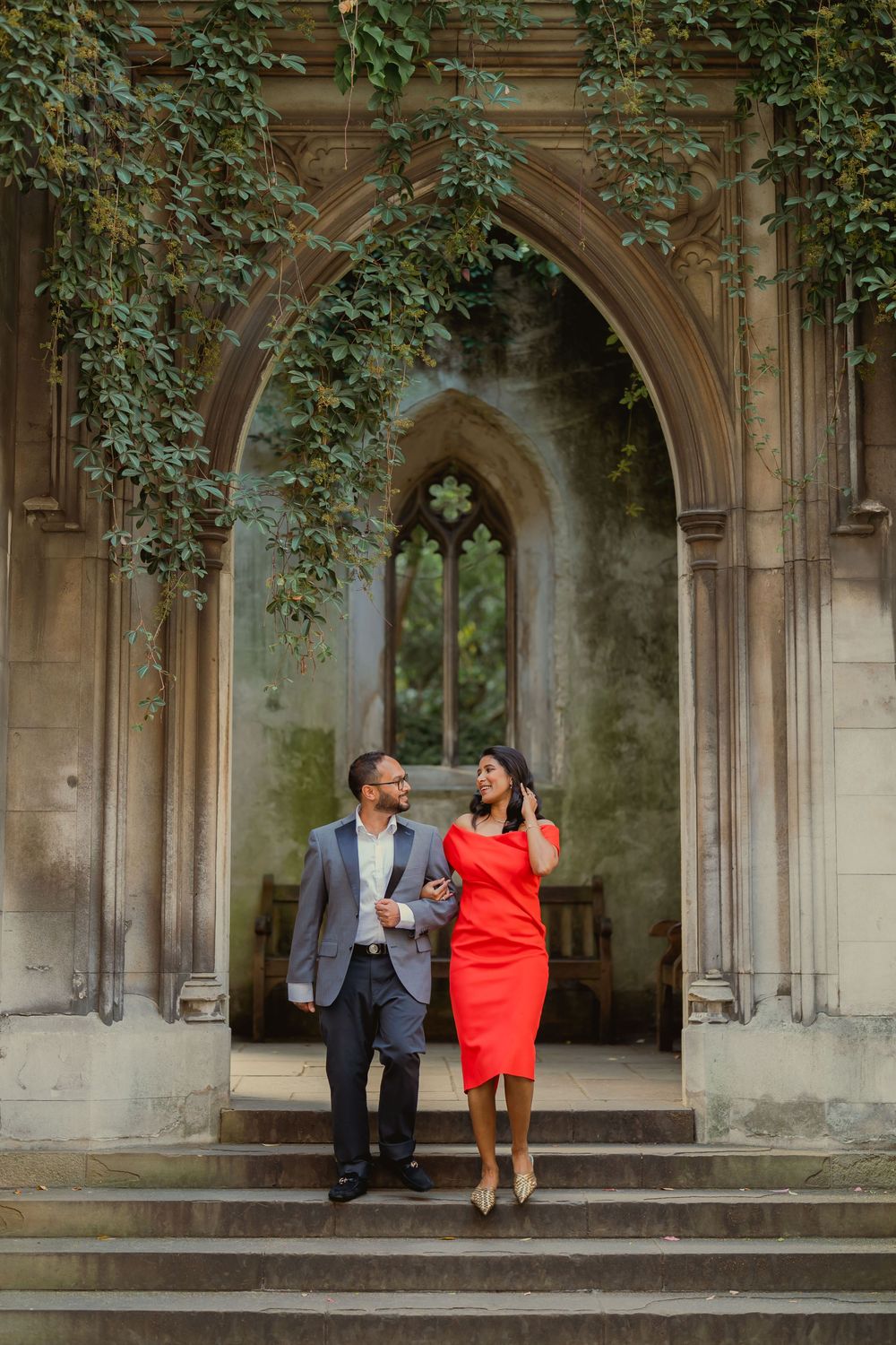 A couple in formal attire walks down stone steps of a Gothic arch building covered in ivy.