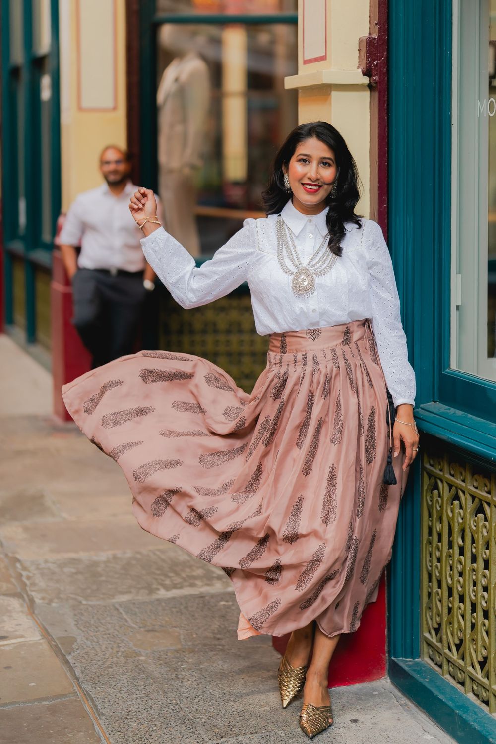 Woman in white blouse and pink printed maxi skirt posing outdoors near colorful storefront.