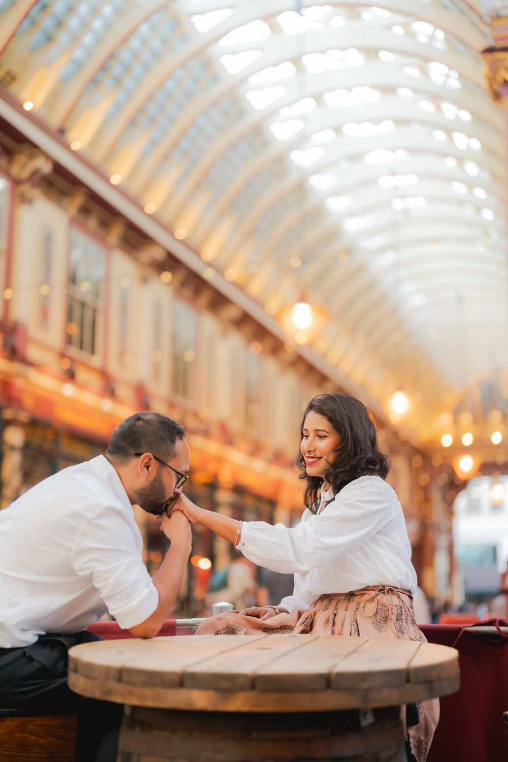 A couple shares a romantic moment at a barrel table inside the elegant Leadenhall Market, London, as he kisses her hand.