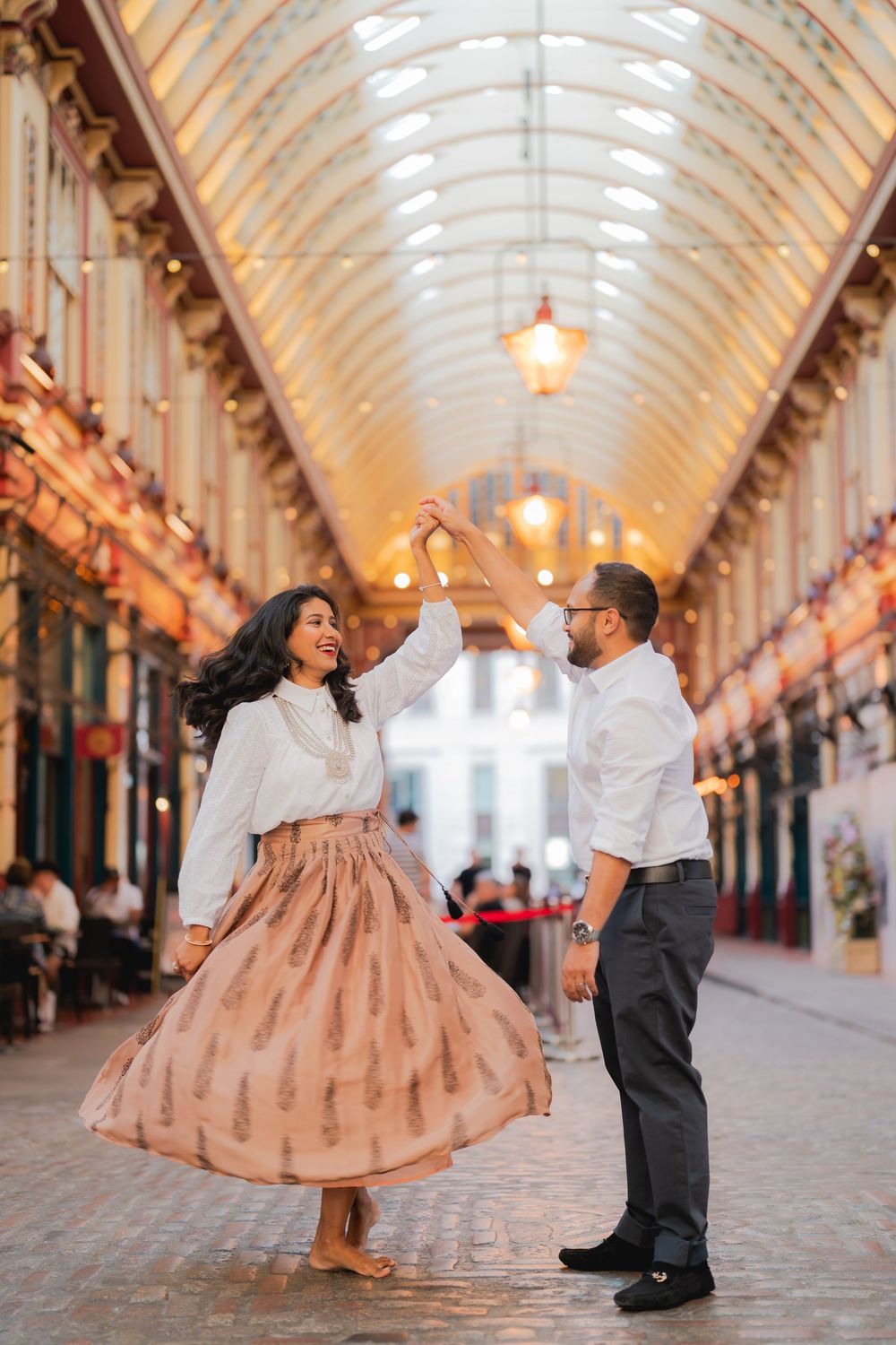 Couple dancing joyfully in Leadenhall Market London, woman in pink skirt twirling under ornate Victorian arcade lights.