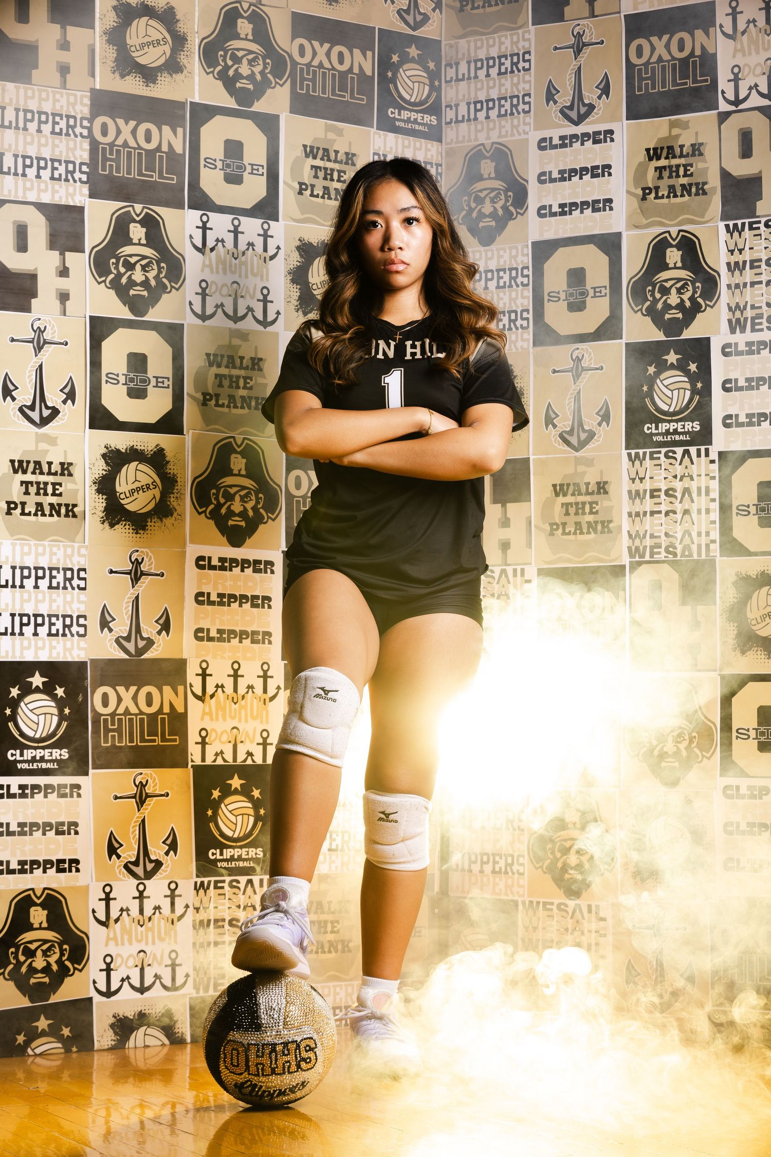 Oxon Hill Clippers volleyball player poses confidently with arms crossed, knee pads, and a volleyball amid dramatic smoke effects.