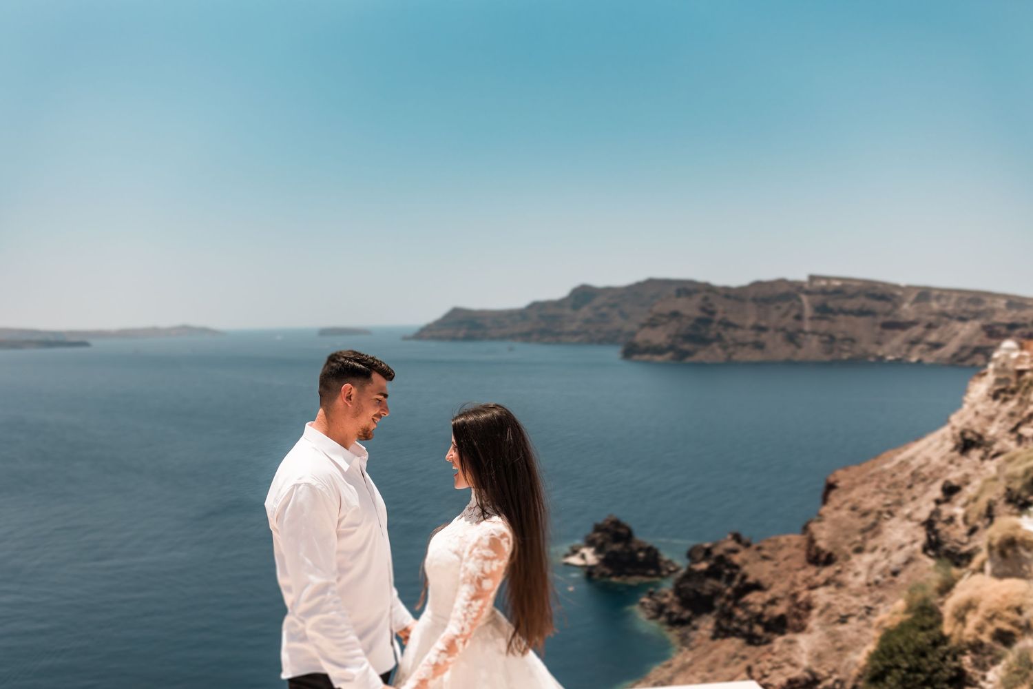 Couple in white attire holds hands facing each other on cliffside overlooking Santorini caldera sea.