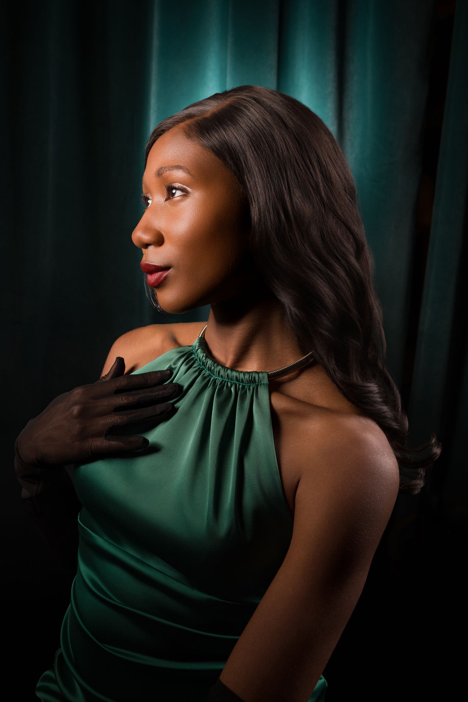 A young Black woman in an elegant green halter dress poses gracefully against a dark teal curtain backdrop.