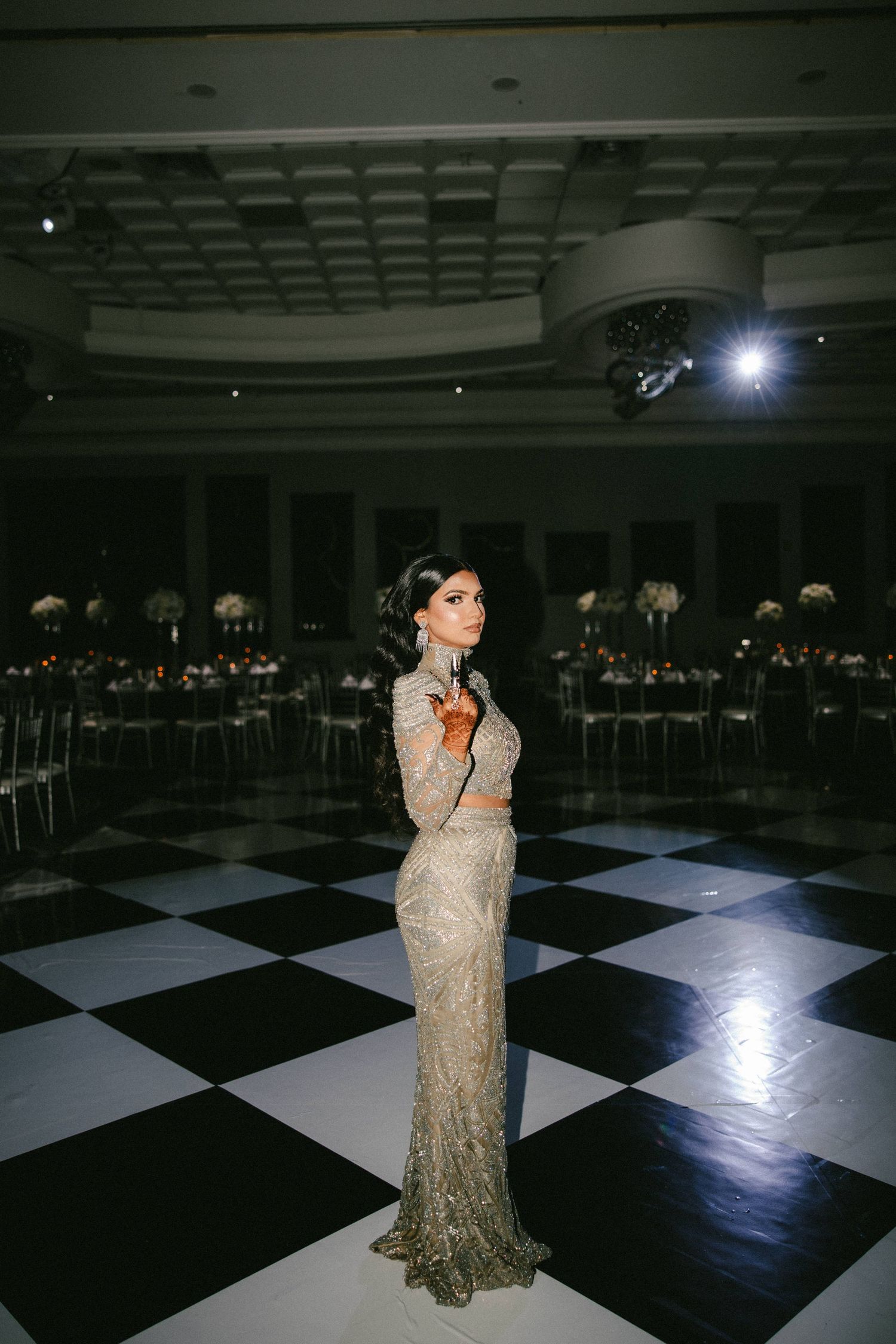 Woman in sequined gown poses on black and white checkered dance floor in elegant ballroom.