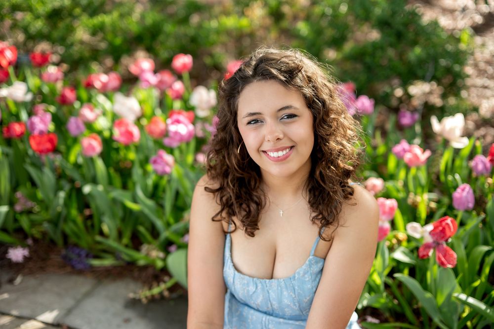 Smiling young woman with curly hair kneeling in front of a vibrant colorful tulip garden in bloom.