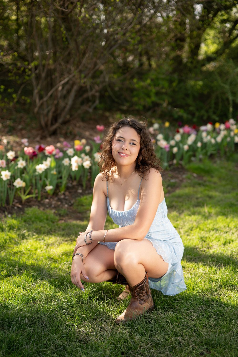 Young woman in blue dress and cowboy boots crouches on grass near colorful tulip garden in sunlight.