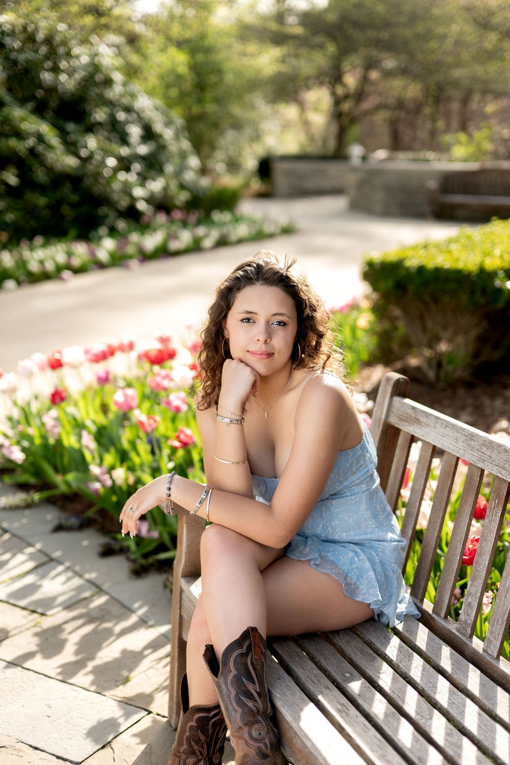 Young woman in blue dress and cowboy boots sits on wooden bench surrounded by spring tulips.
