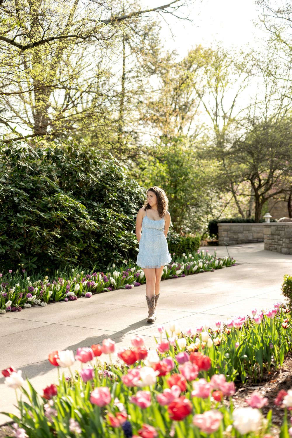 Young woman in blue dress and cowboy boots strolls through a vibrant tulip garden on a sunny day.