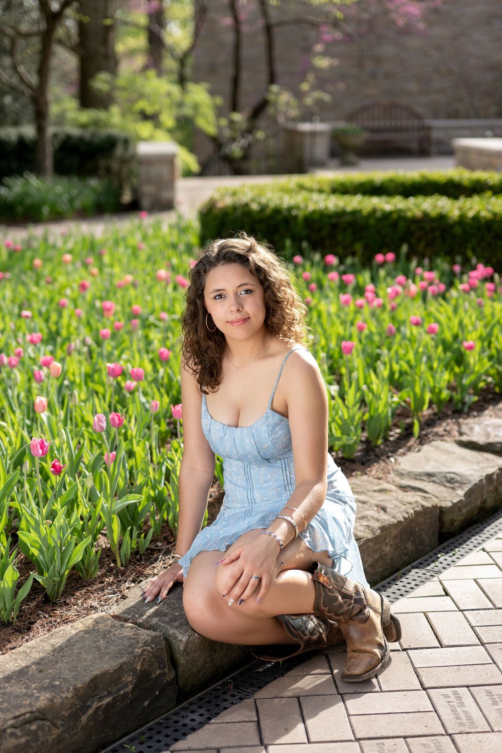 Young woman in blue dress and cowboy boots sits on garden border surrounded by bright pink tulips.