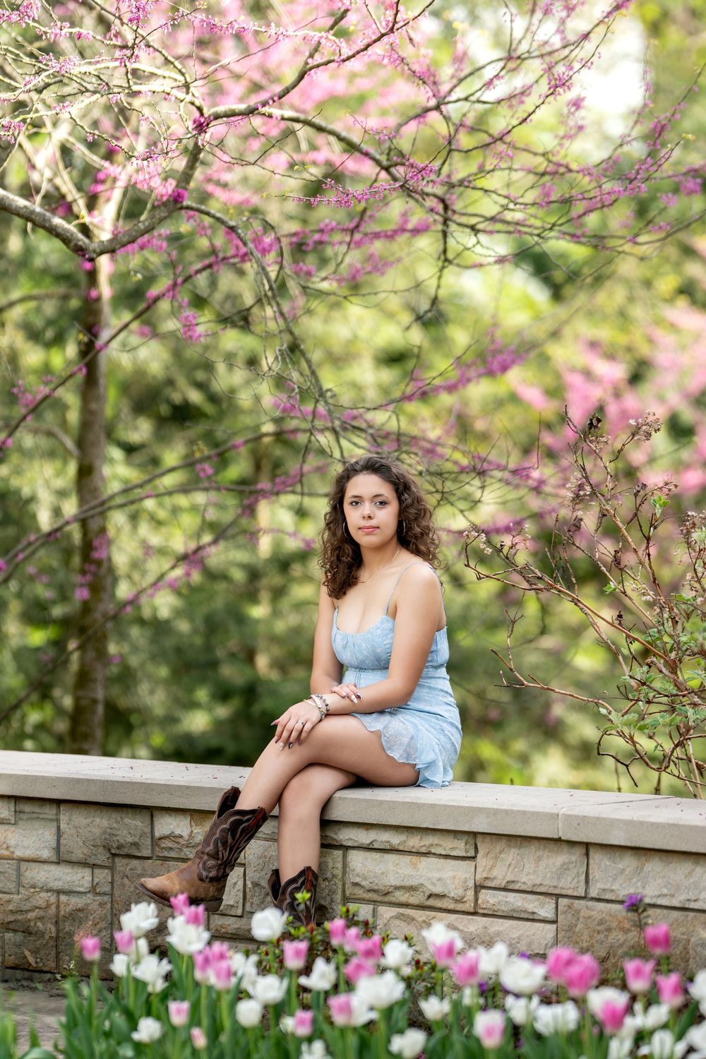 Young woman in blue dress and cowboy boots sitting on stone wall with pink cherry blossoms.