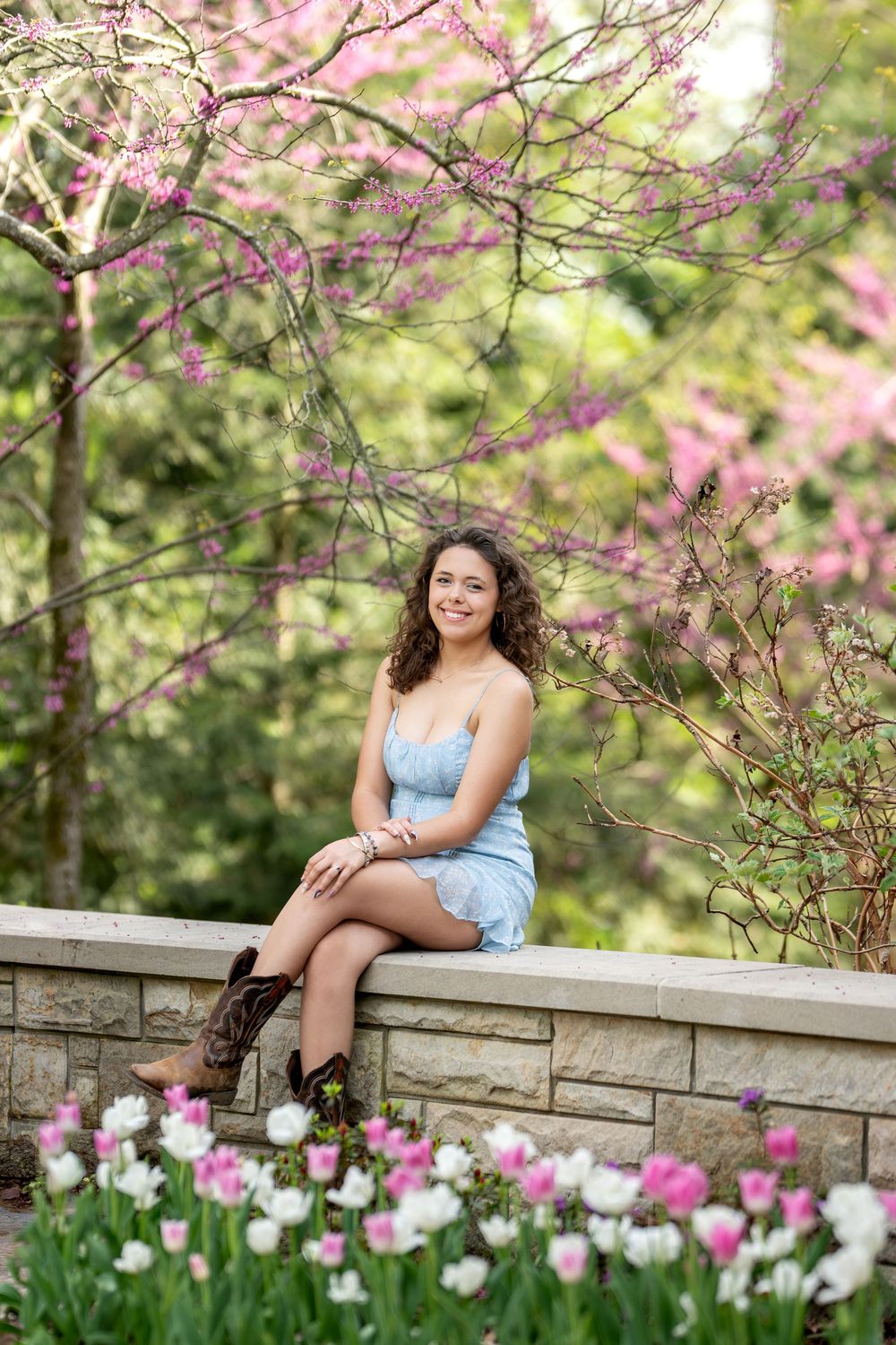 Smiling young woman in light blue dress seated on stone ledge with tulips and pink blossoms.