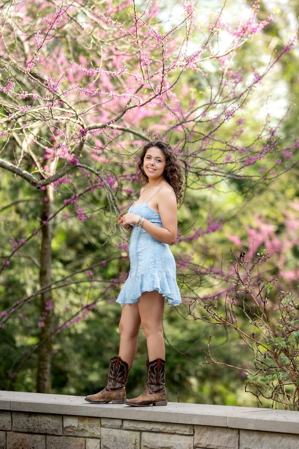 Young woman in blue dress and cowboy boots standing on stone wall under pink flowering tree.