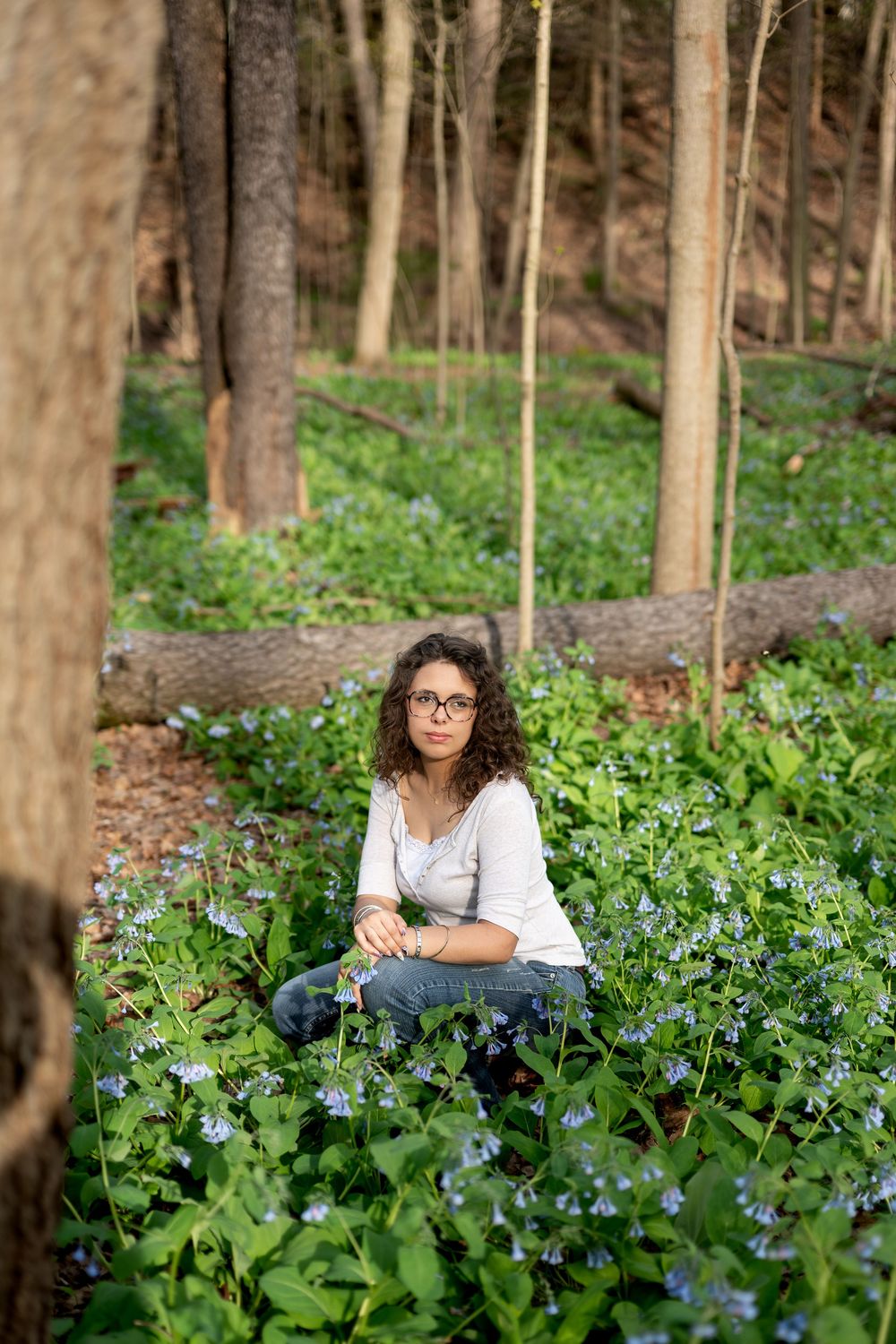 Young woman with glasses sitting among blue Virginia bluebells in a sunlit woodland forest.
