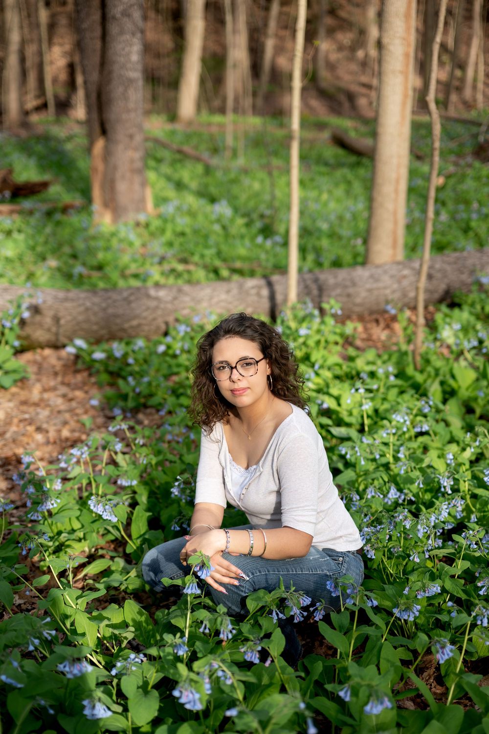 Teen girl in glasses and white top kneeling among blue wildflowers in a spring forest setting.