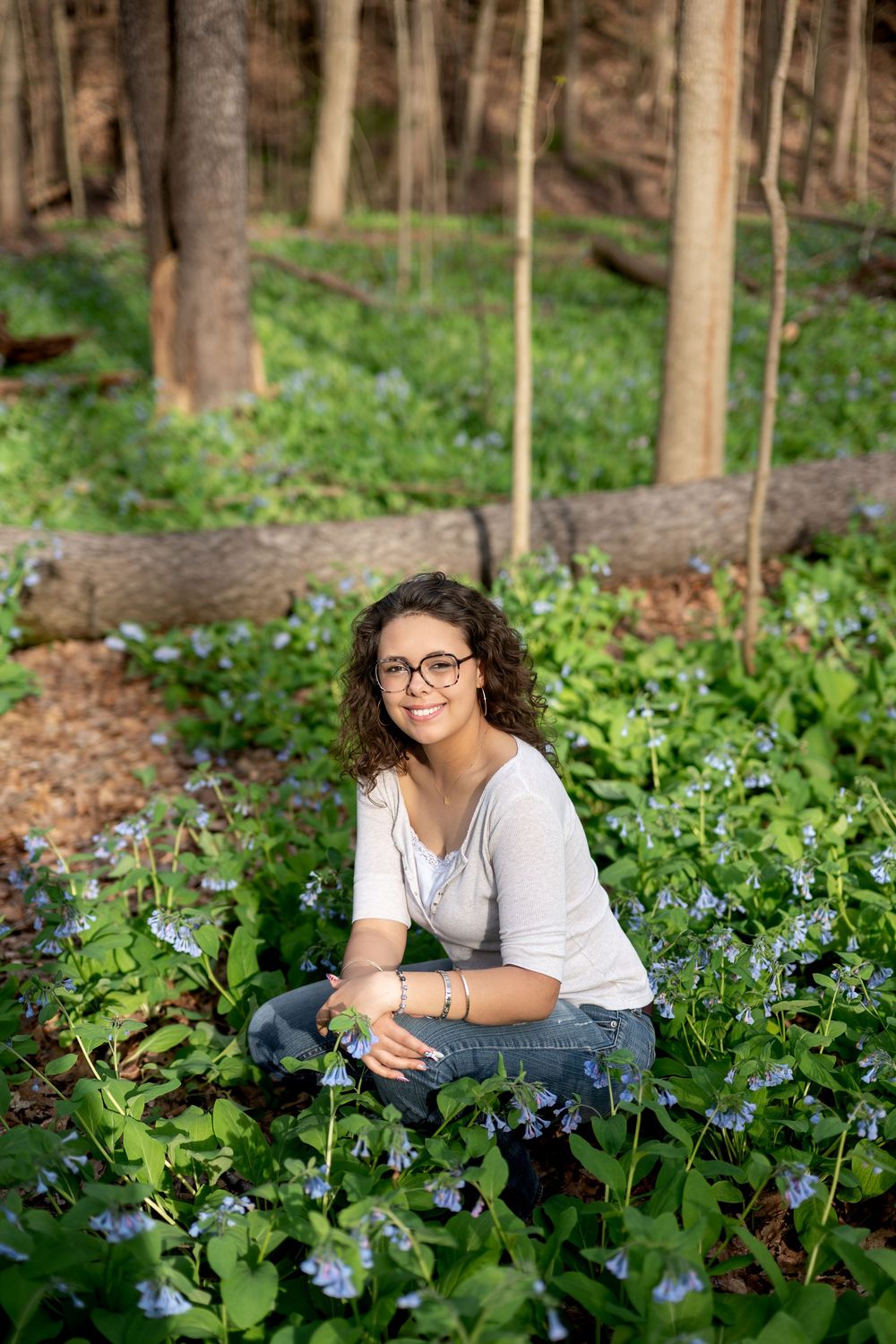 Smiling young woman with glasses posing among blue wildflowers on a forest floor in spring.