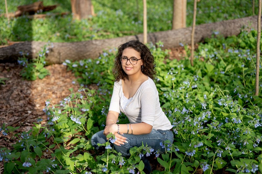 Woman with curly hair and glasses sitting among blue wildflowers in a woodland garden.