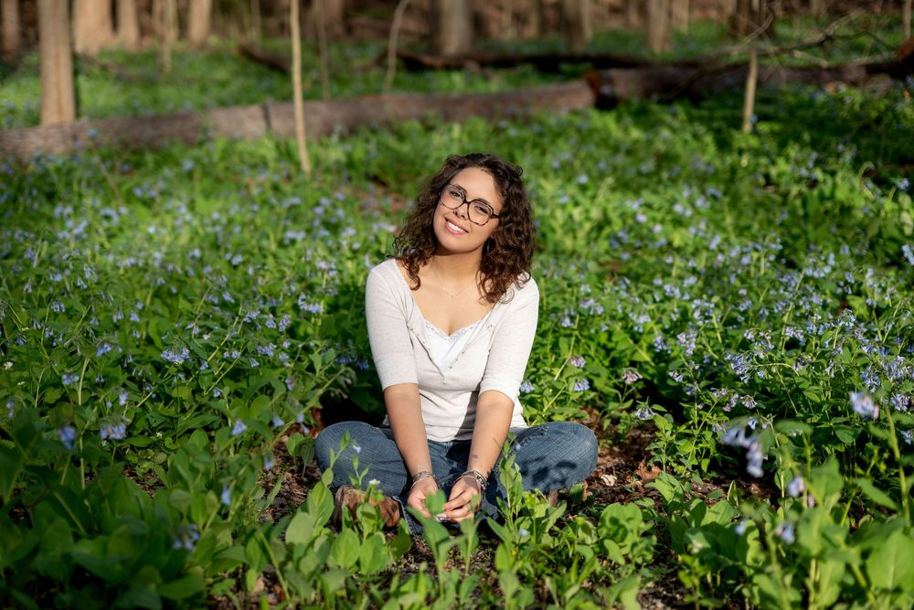 Curly-haired woman in white top sitting peacefully among blue wildflowers in a sunlit forest.
