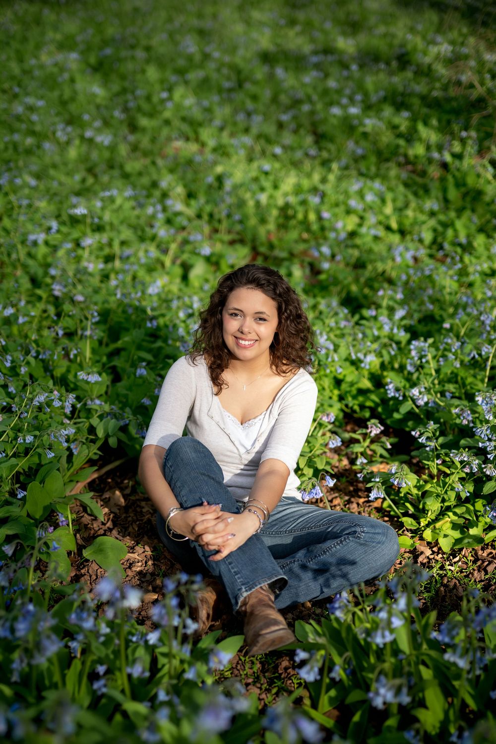Smiling woman in white top seated among vibrant blue wildflowers in a green woodland setting.