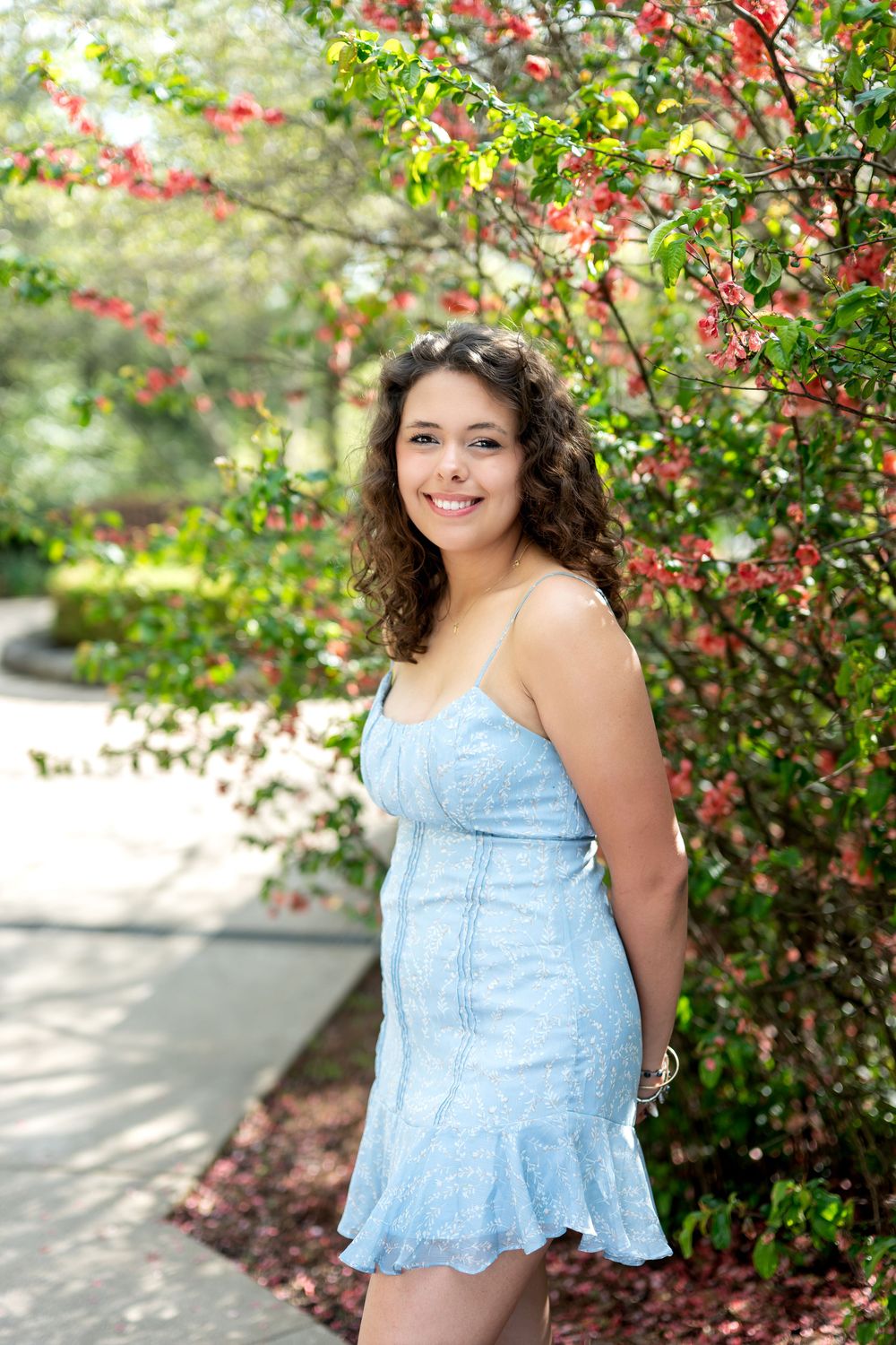 Smiling young woman in a light blue sundress standing beside vibrant pink flowering bushes in a garden.